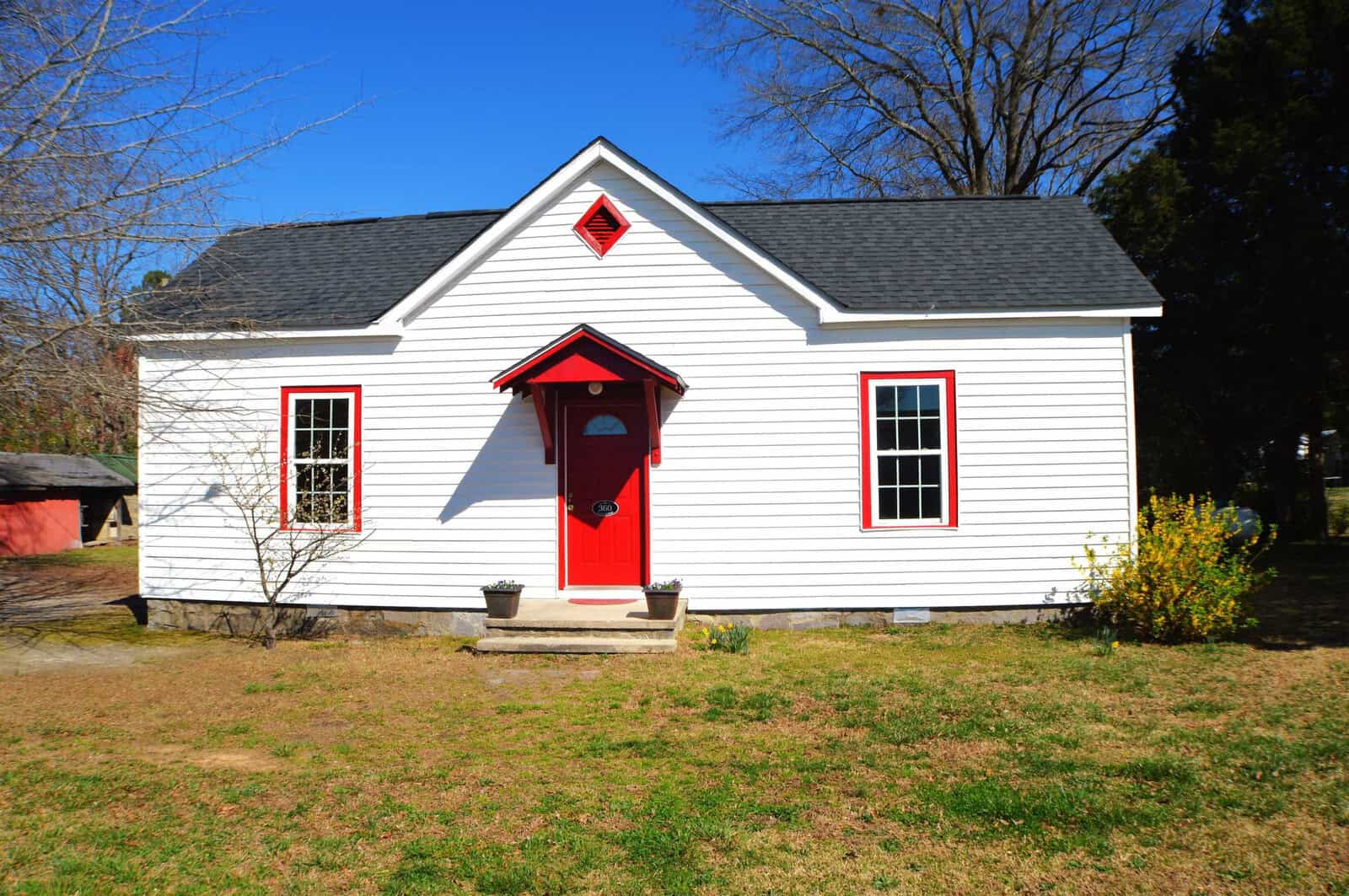 A small white house with red trim and a red door, situated under a clear blue sky.