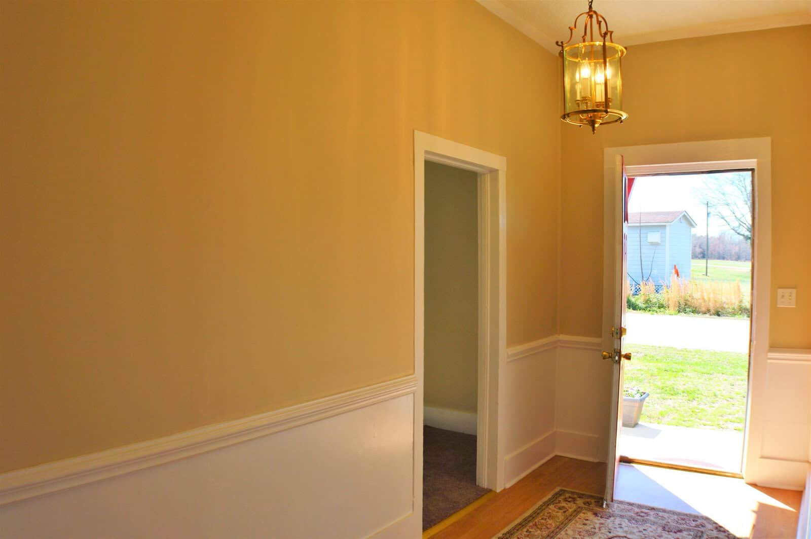 A warmly lit interior hallway with an open door leading to a bright outdoor space, featuring a hanging lantern light fixture and white wainscoting on the lower wall.