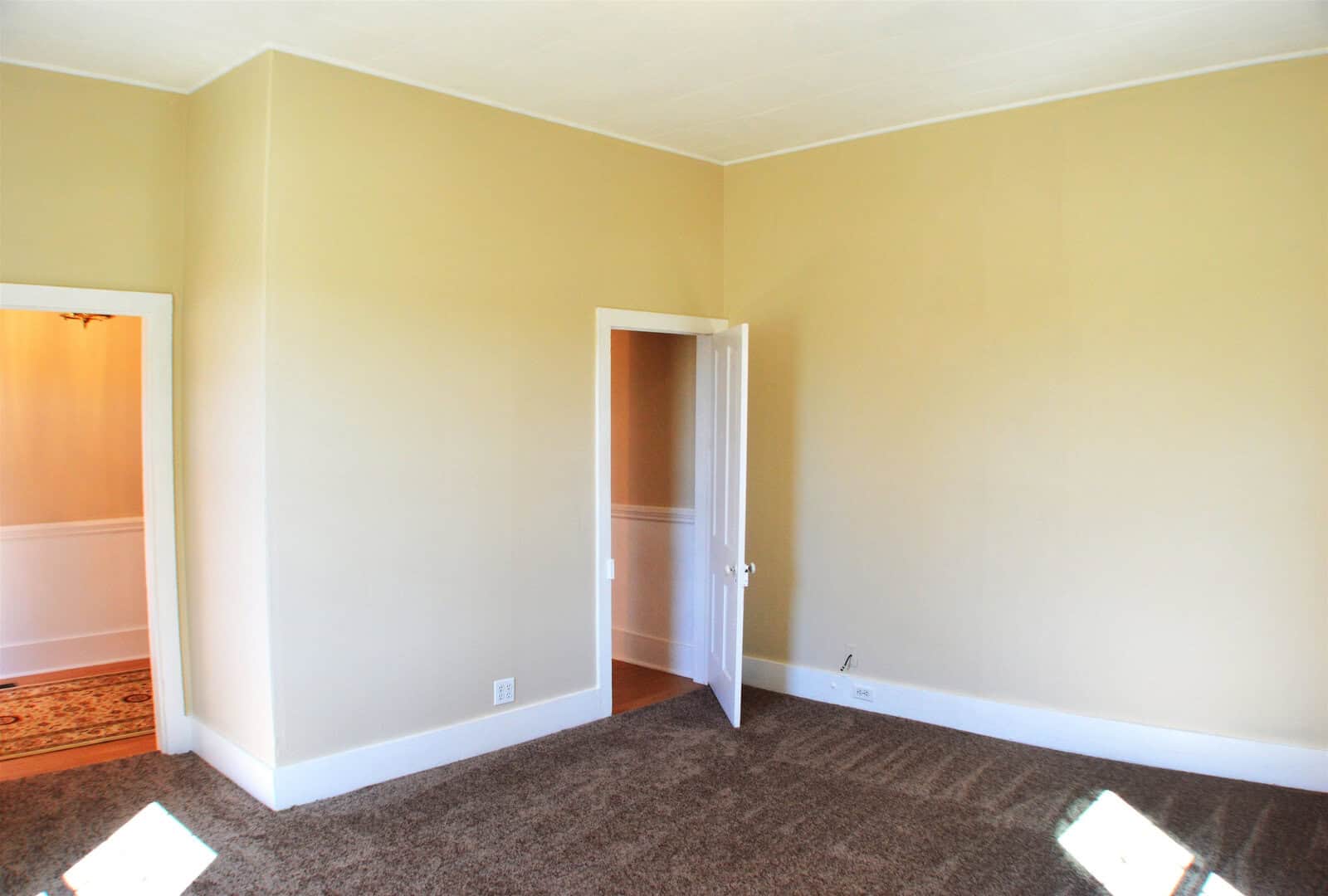 A well-lit empty room with beige walls, brown carpet, and white trim featuring an open door leading to an adjoining room.