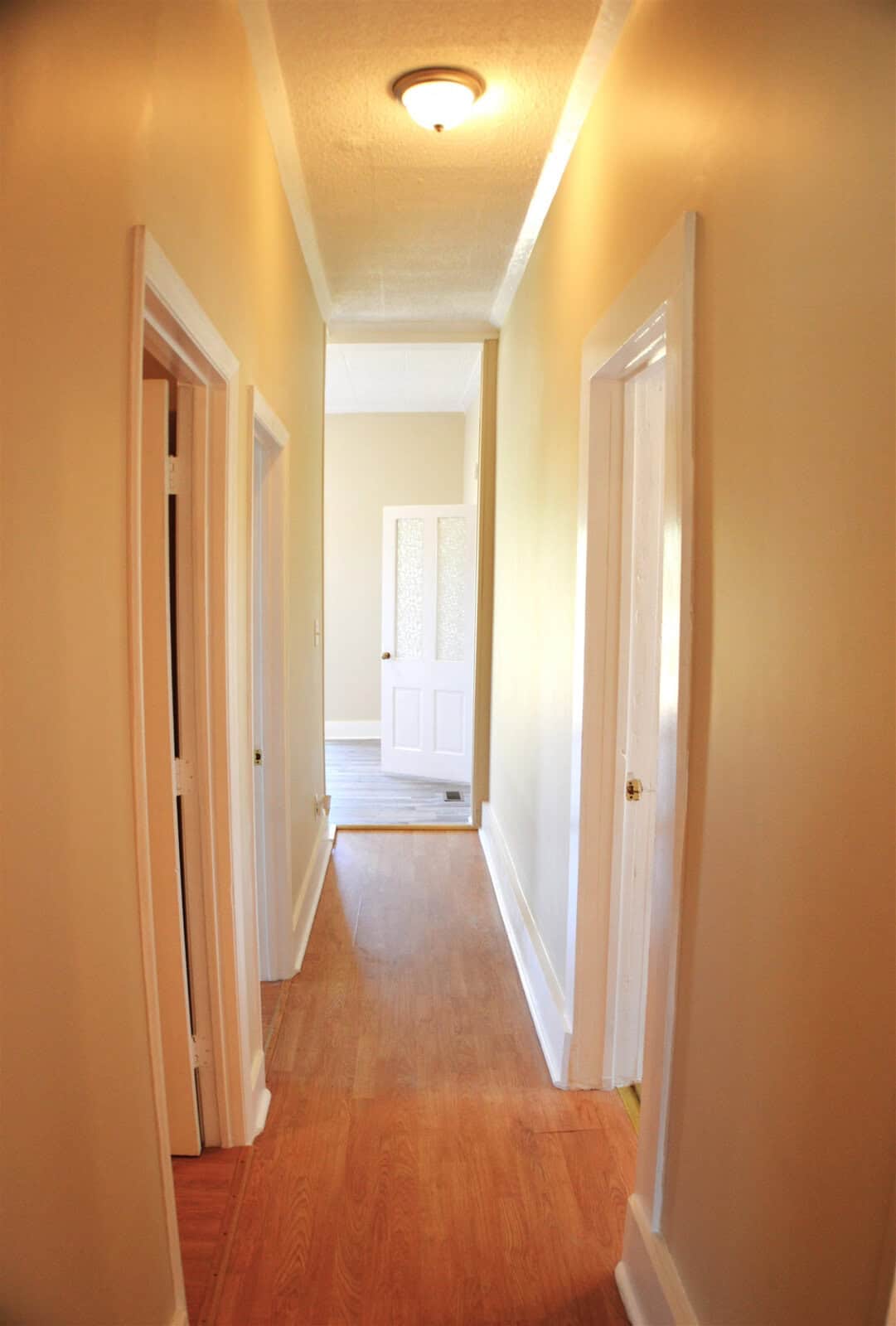 A brightly lit, narrow hallway with wooden floors, white walls, and multiple white doors leading to different rooms.