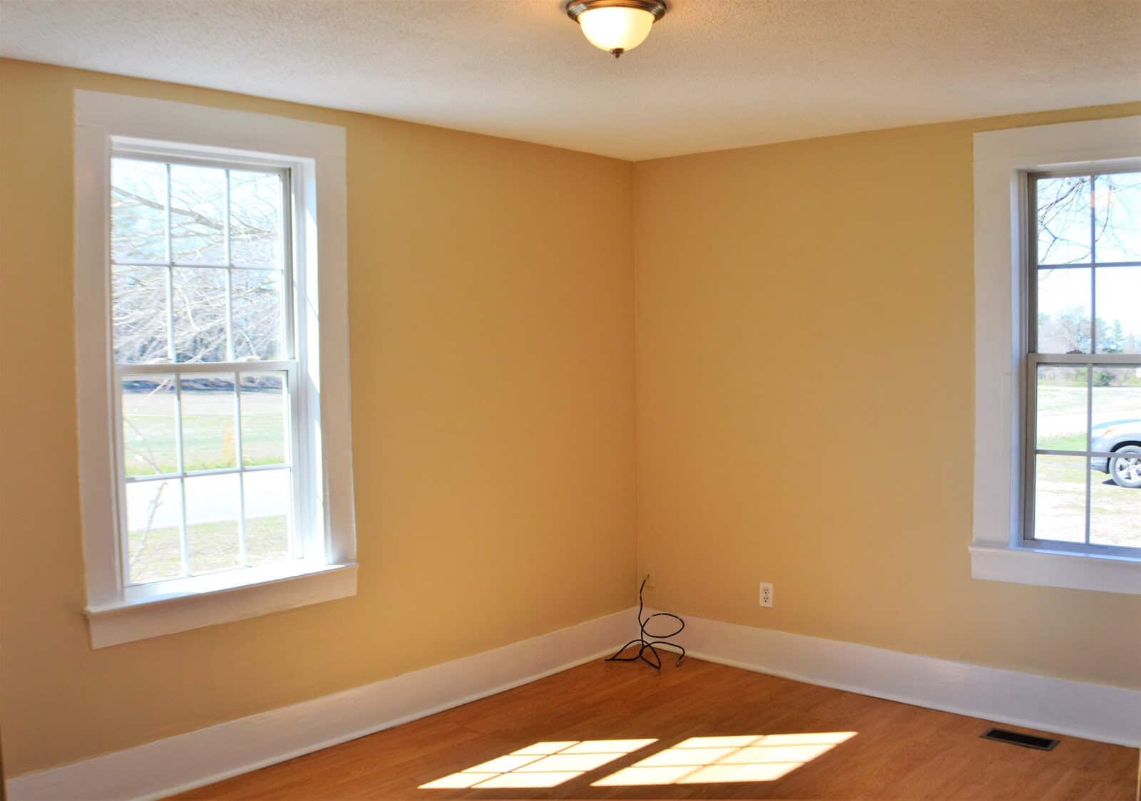 An empty room with beige walls and hardwood flooring featuring two windows that provide a view of the outdoors and allow natural light to enter the space. there is a single light fixture on the ceiling and an electrical cord on the floor.