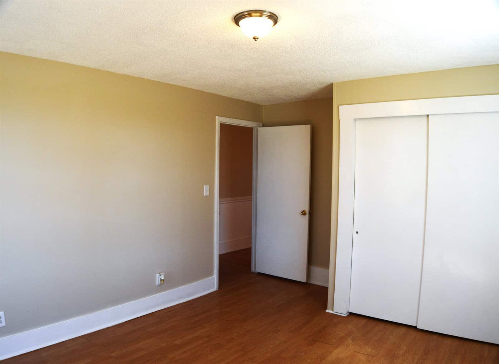 An empty room with beige walls, a wooden floor, a closed white door, and a white closed closet.