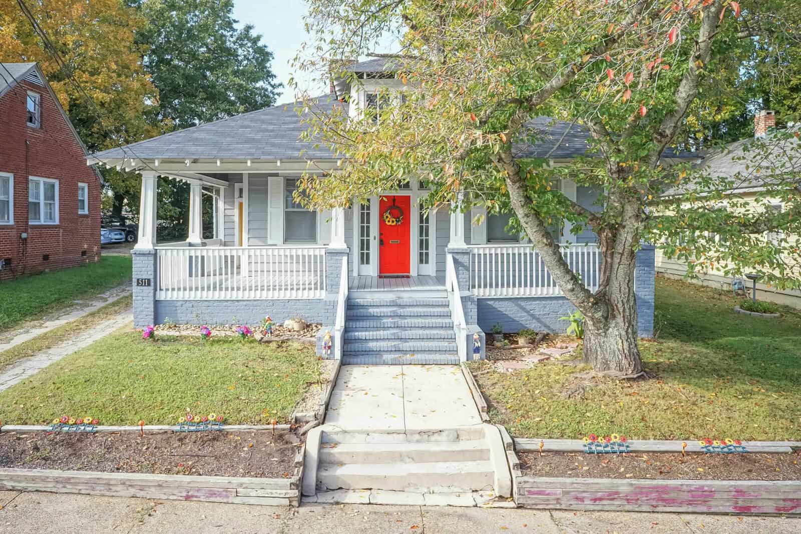 A single-story house with a red door and front porch, surrounded by greenery and a clear sky above.