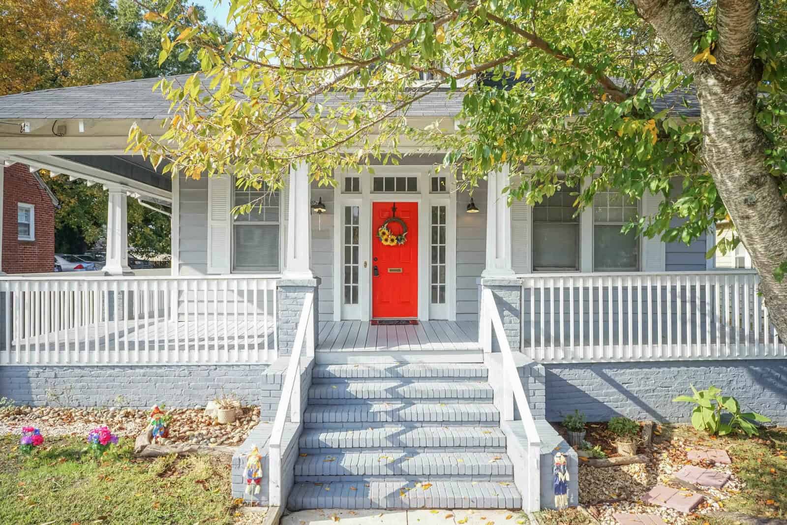 A single-story house with a gray exterior, white trim, and a bold red front door, flanked by two small shrubs and featuring a covered front porch with steps leading up to it, all under the shade of a large tree.