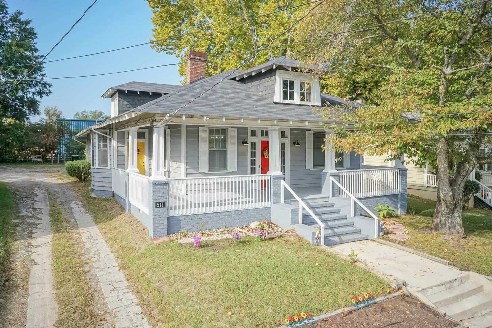 A single-story gray house with white trim, a red front door, and a small front porch surrounded by a low white fence, situated in a neighborhood with trees and a clear sky.