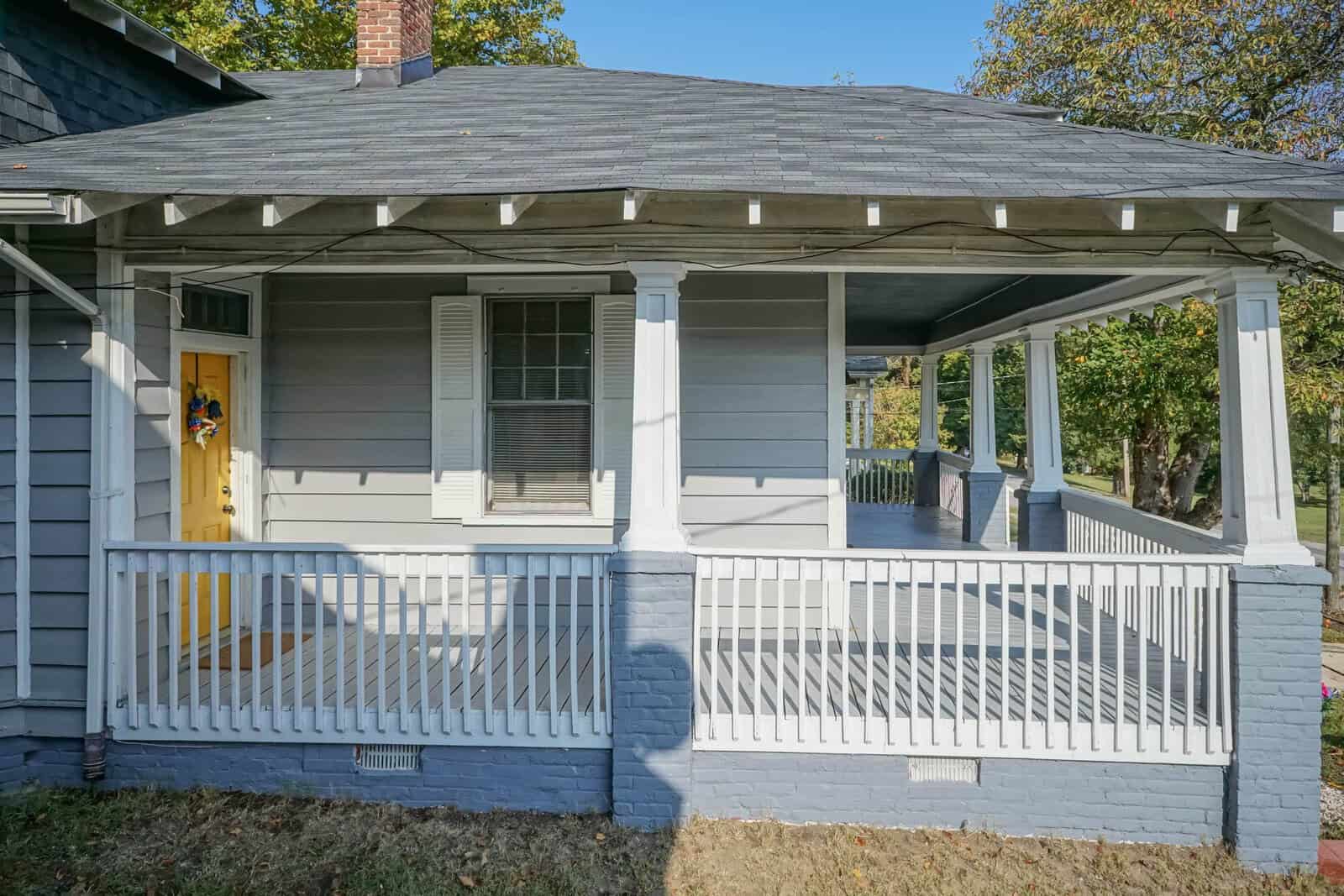 A single-story gray house with white trim, featuring a covered front porch with a white railing and a yellow front door decorated with a wreath.