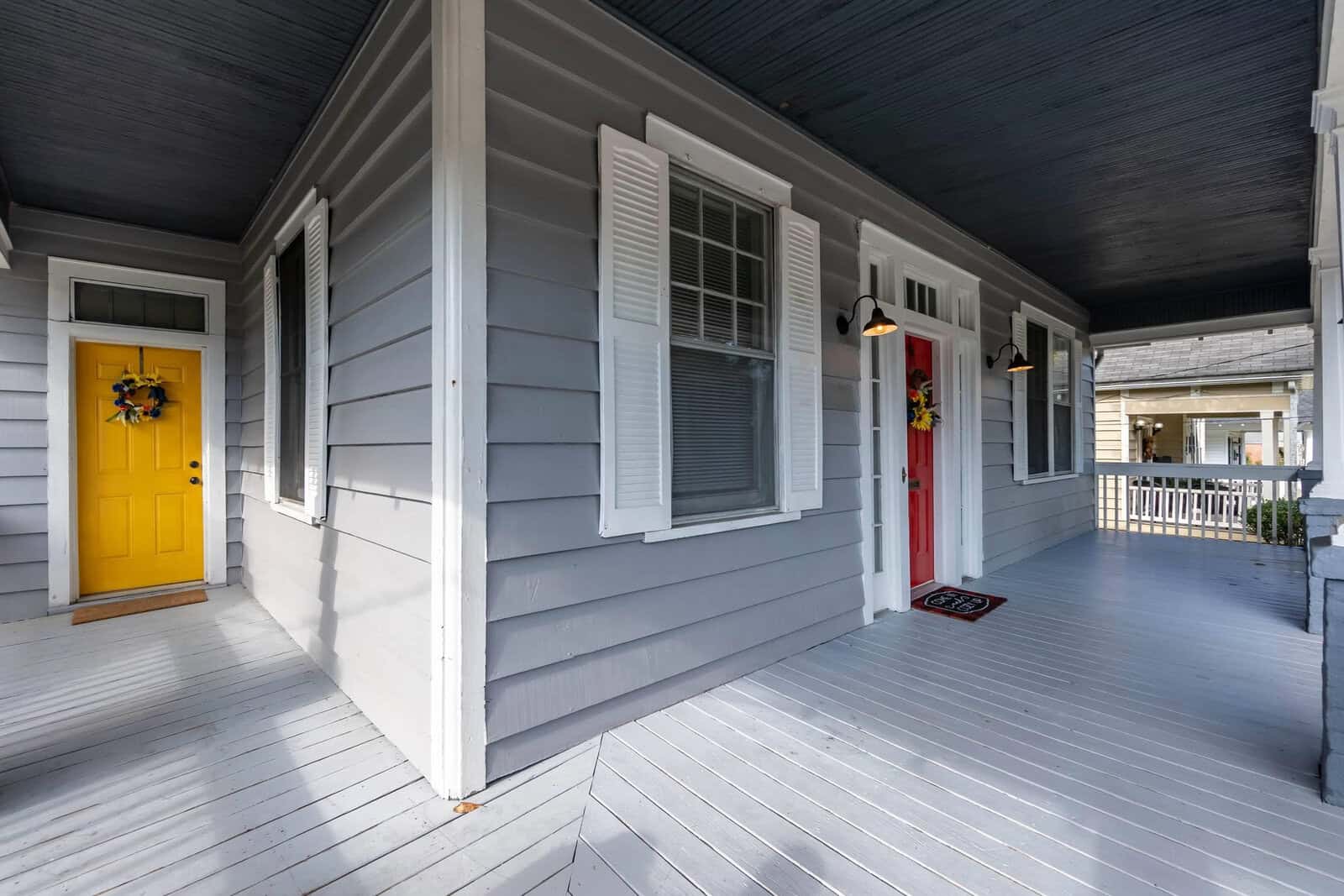 A front porch of a house featuring a bright yellow door, flanked by white windows with black shutters, and decorated with wreaths.
