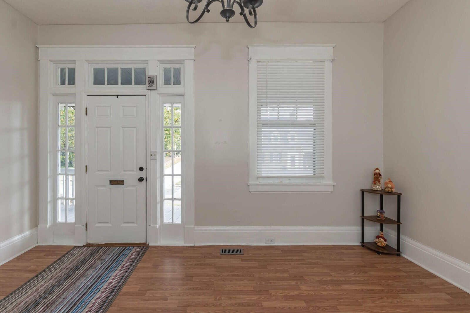 An empty room featuring a white front door with sidelights, a window with blinds, wooden flooring, and a small shelf with decorative items in the corner.