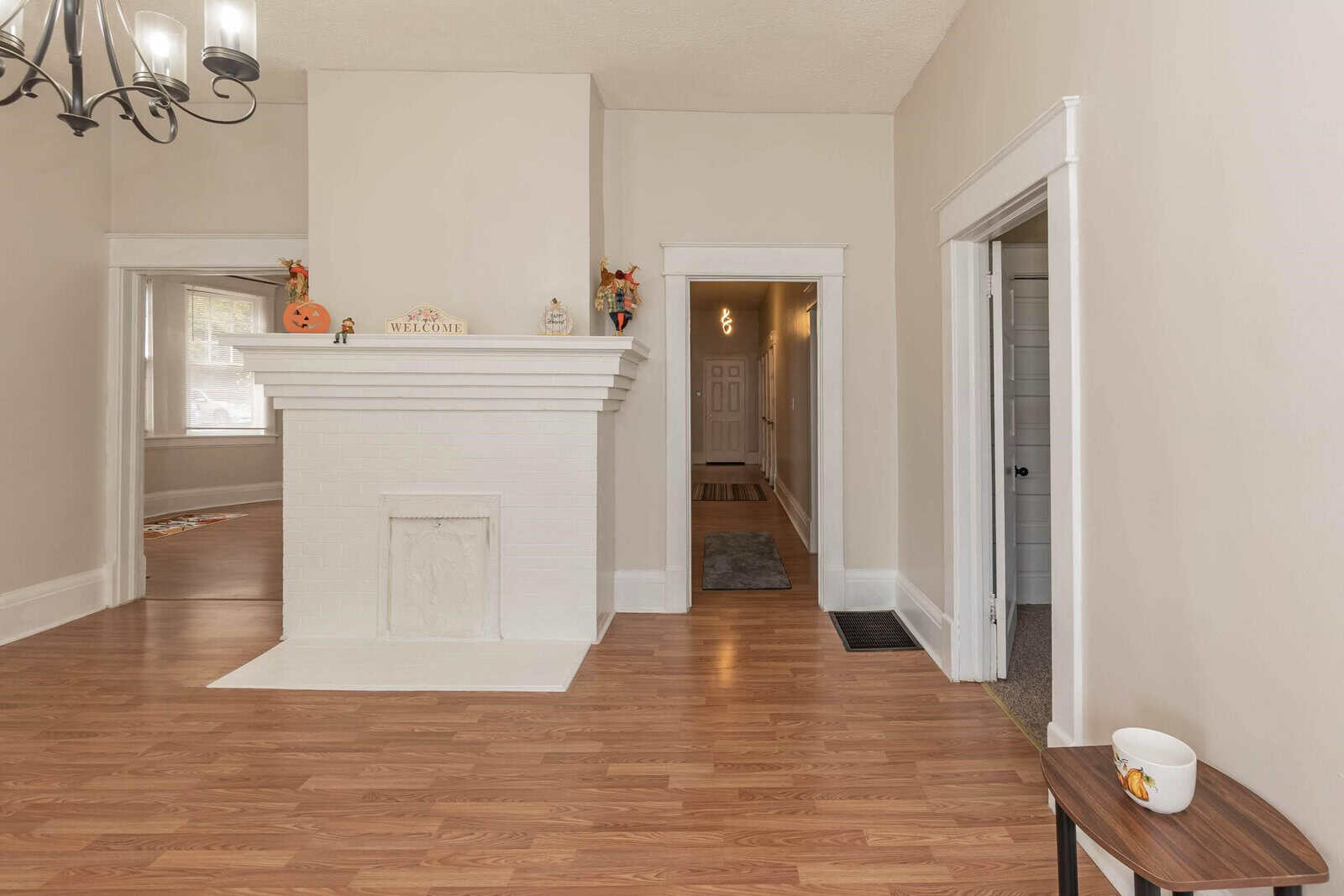 A view of a well-lit, empty living room with a sealed fireplace and autumnal decorations on the mantel. the room features hardwood flooring, white walls, and an open doorway leading to another part of the house.