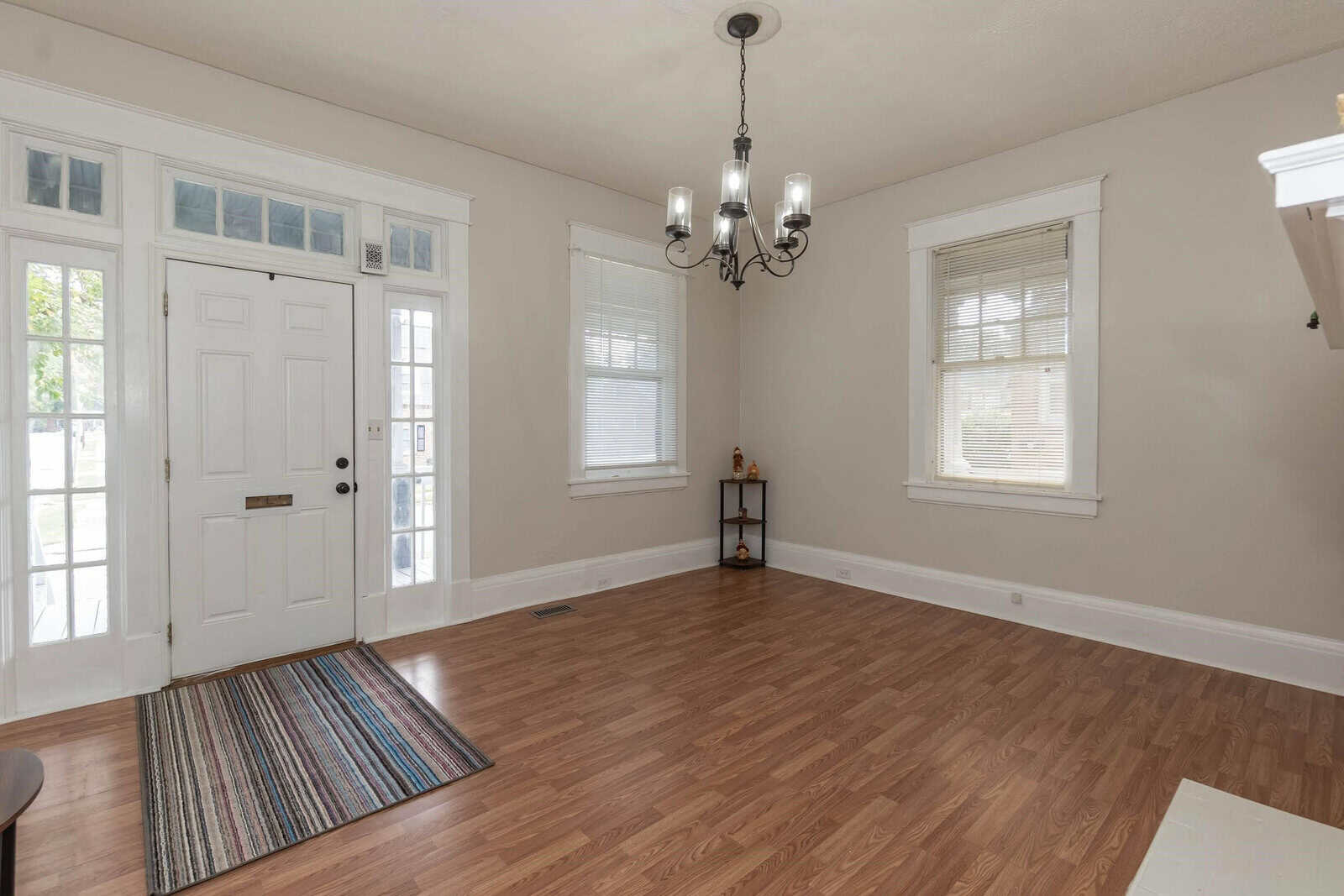 An empty room with hardwood floors, a white front door with sidelights, two windows draped with blinds, and a small wooden shelving unit in the corner. a multicolored rug is placed in front of the door, and a traditional chandelier hangs from the ceiling.