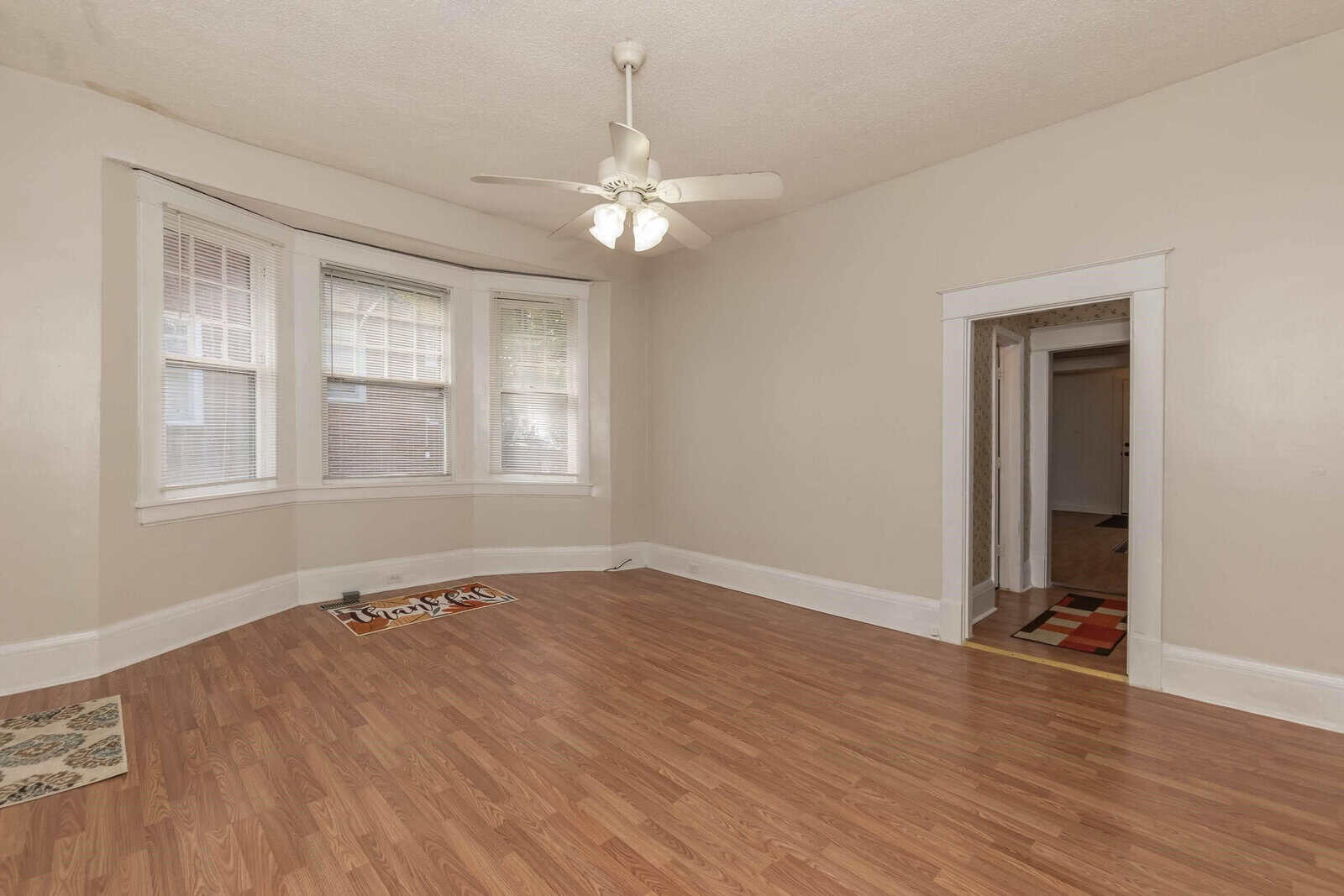 An empty room with beige walls, laminate flooring, two windows with blinds, a ceiling fan with lights, and an open doorway.