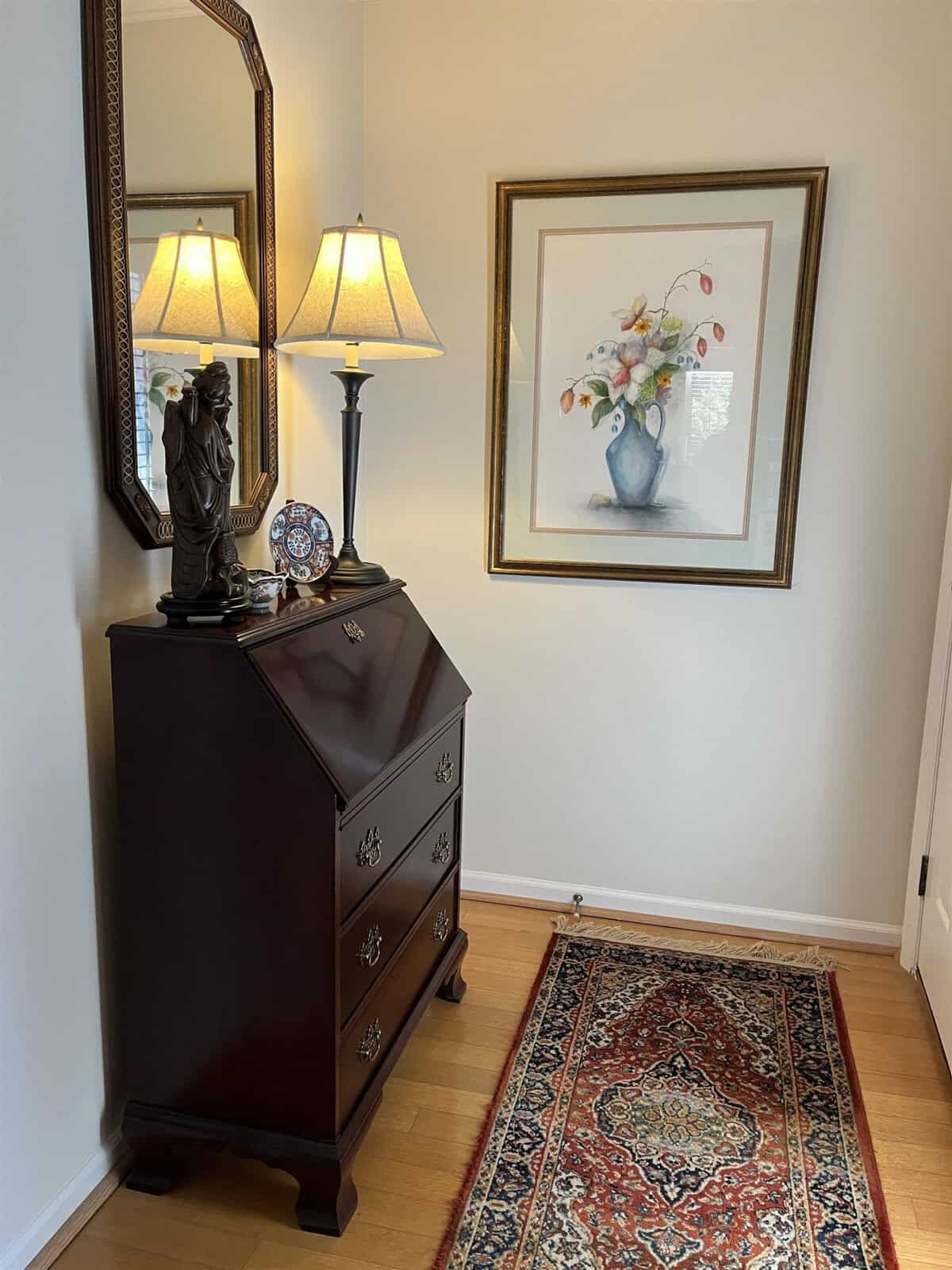 A well-appointed corner of a room featuring a wooden chest of drawers with a lamp and a small statue atop it, a framed floral artwork on the wall, a decorative mirror, and a persian rug on the hardwood floor.