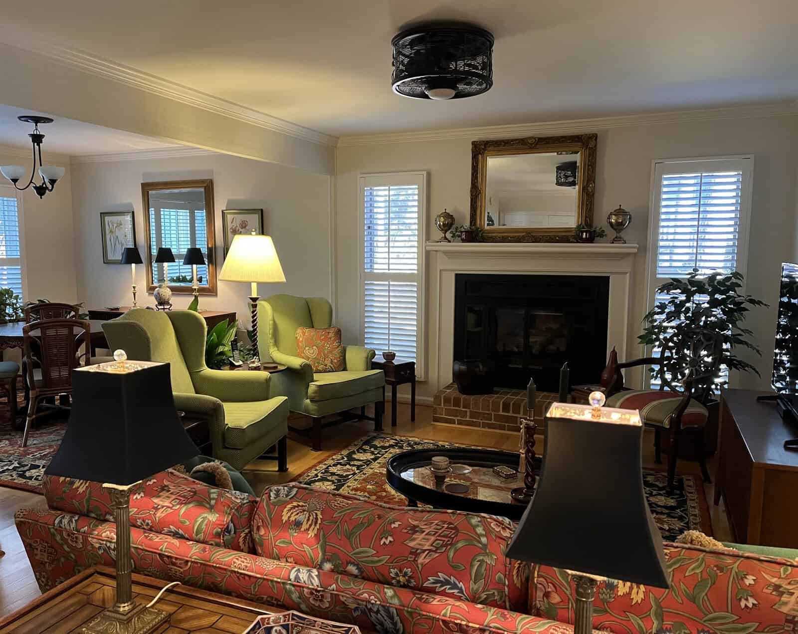 A well-lit, cozy living room with a fireplace, furnished with patterned sofas, green armchairs, lamps, and a chandelier.