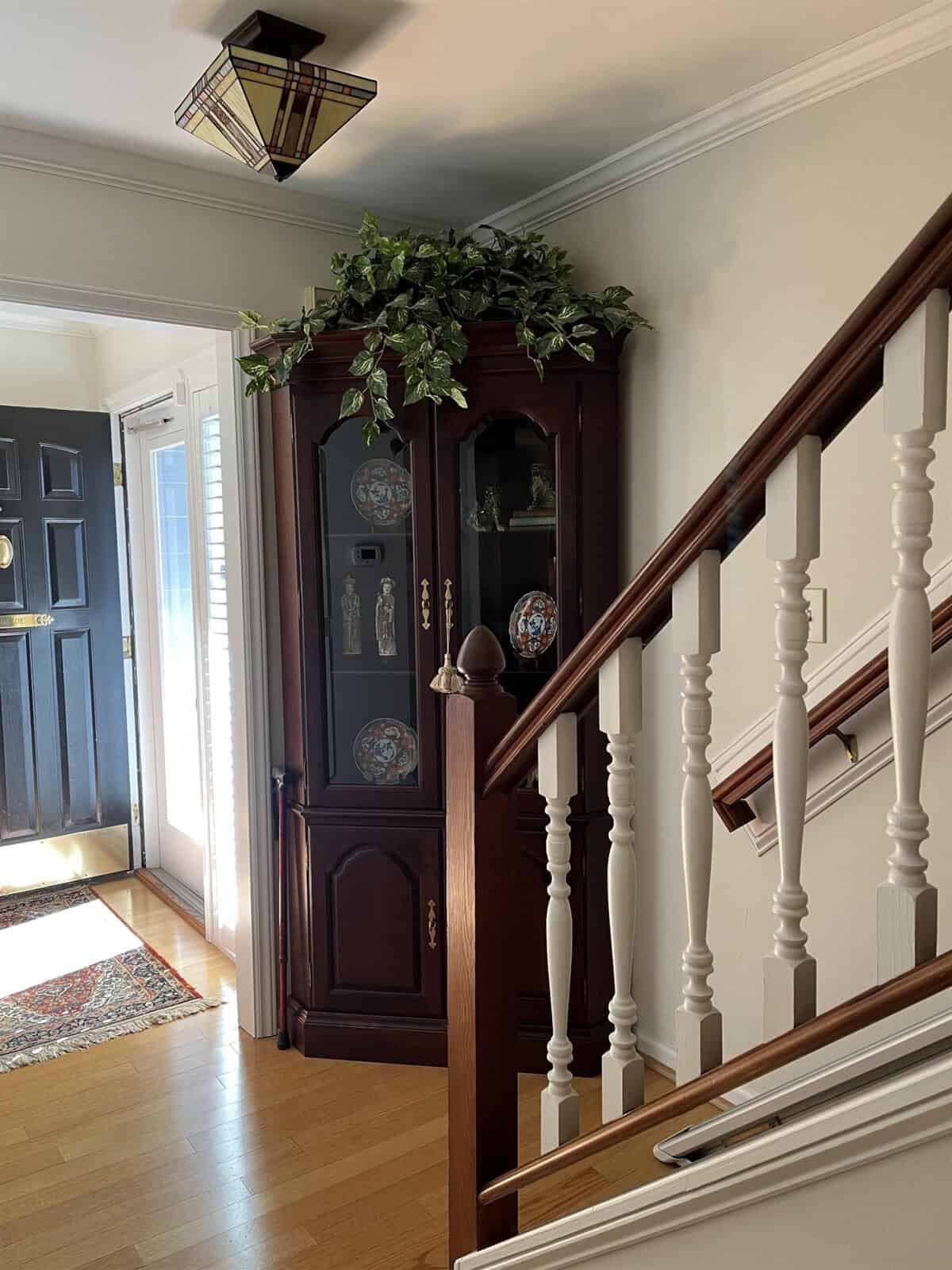 An elegant entryway featuring a wooden staircase with white balusters, a dark-toned corner curio cabinet adorned with decorative plates and topped with greenery, complemented by warm lighting and a traditional rug.