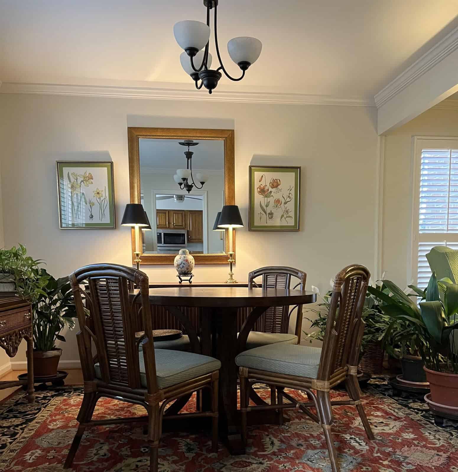 A well-lit dining room with a wooden table, four chairs, decorative lamps, and framed artwork on the walls, reflecting a contrasting view in a large mirror.