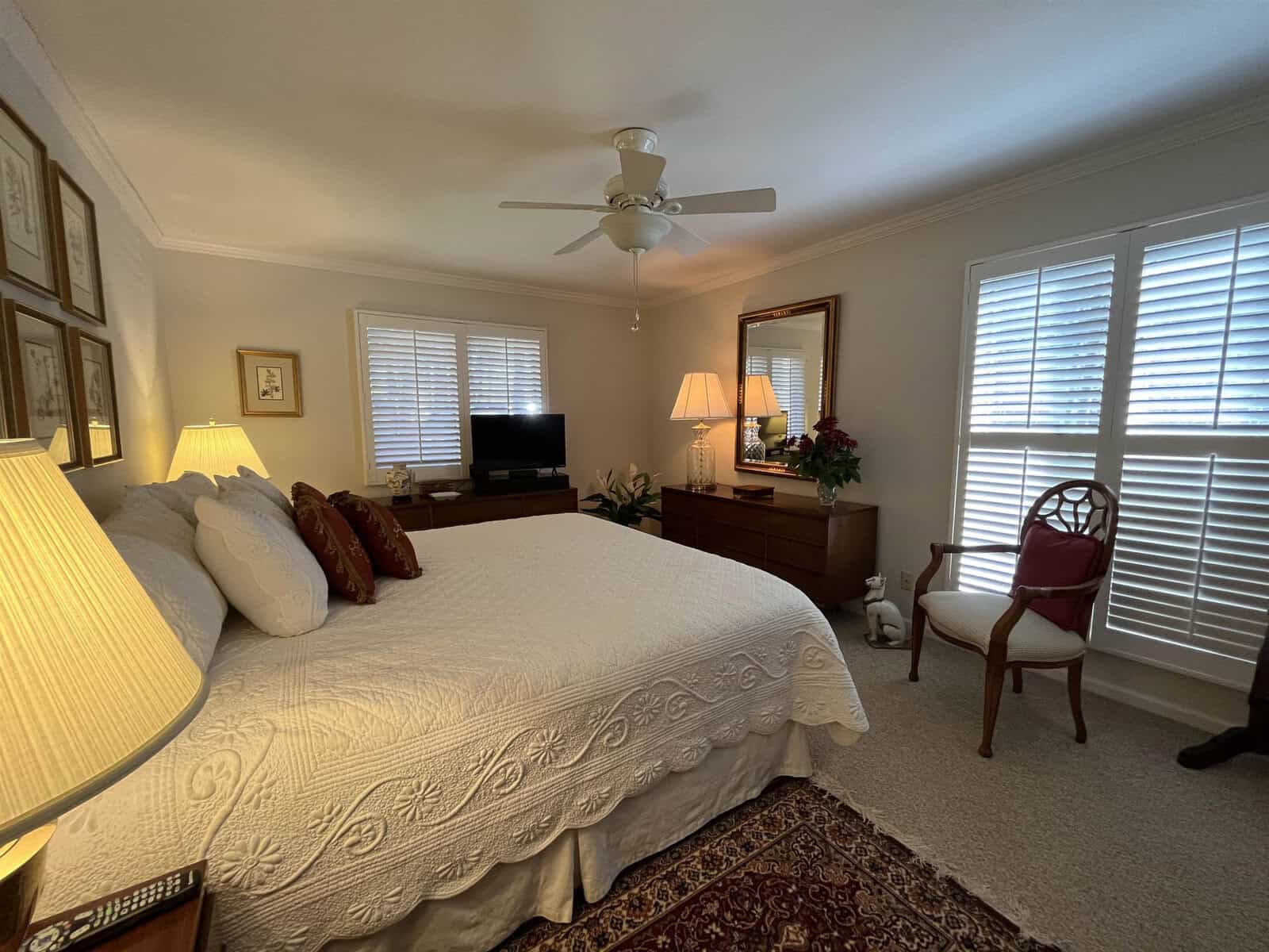 A well-lit, tidy bedroom featuring a large bed with decorative pillows, two nightstands with lamps, a ceiling fan, a television, framed wall art, and a chair by the window.