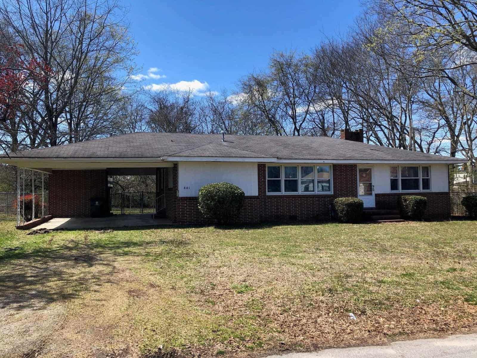 A single-story brick house with a large front yard and bare trees in the background on a sunny day.