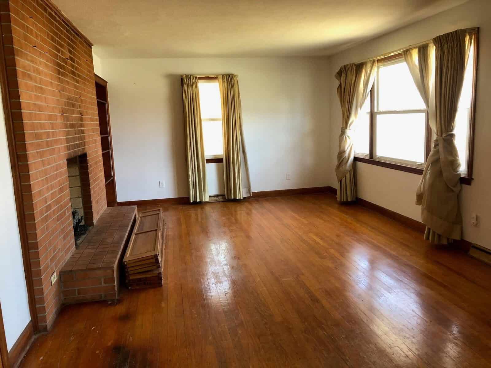 An empty room with hardwood floors, brick fireplace, and draped curtains on windows.