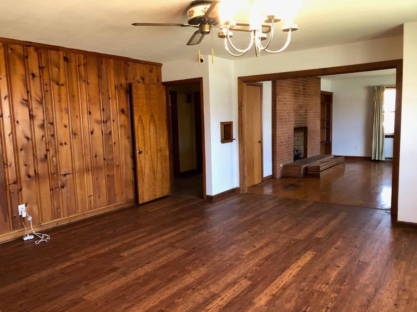 A spacious room with hardwood floors featuring a ceiling fan and chandelier, wooden cabinetry, and brick fireplace, leading into an adjacent room.