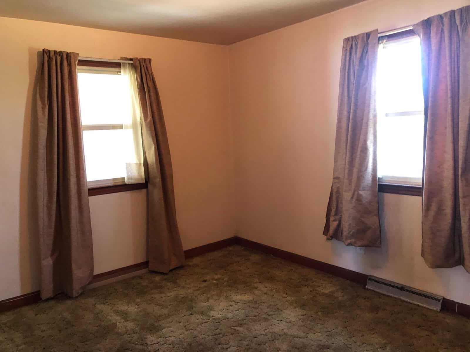 A sparsely furnished room with peach-colored walls, two windows with brown curtains, and a brown carpeted floor.