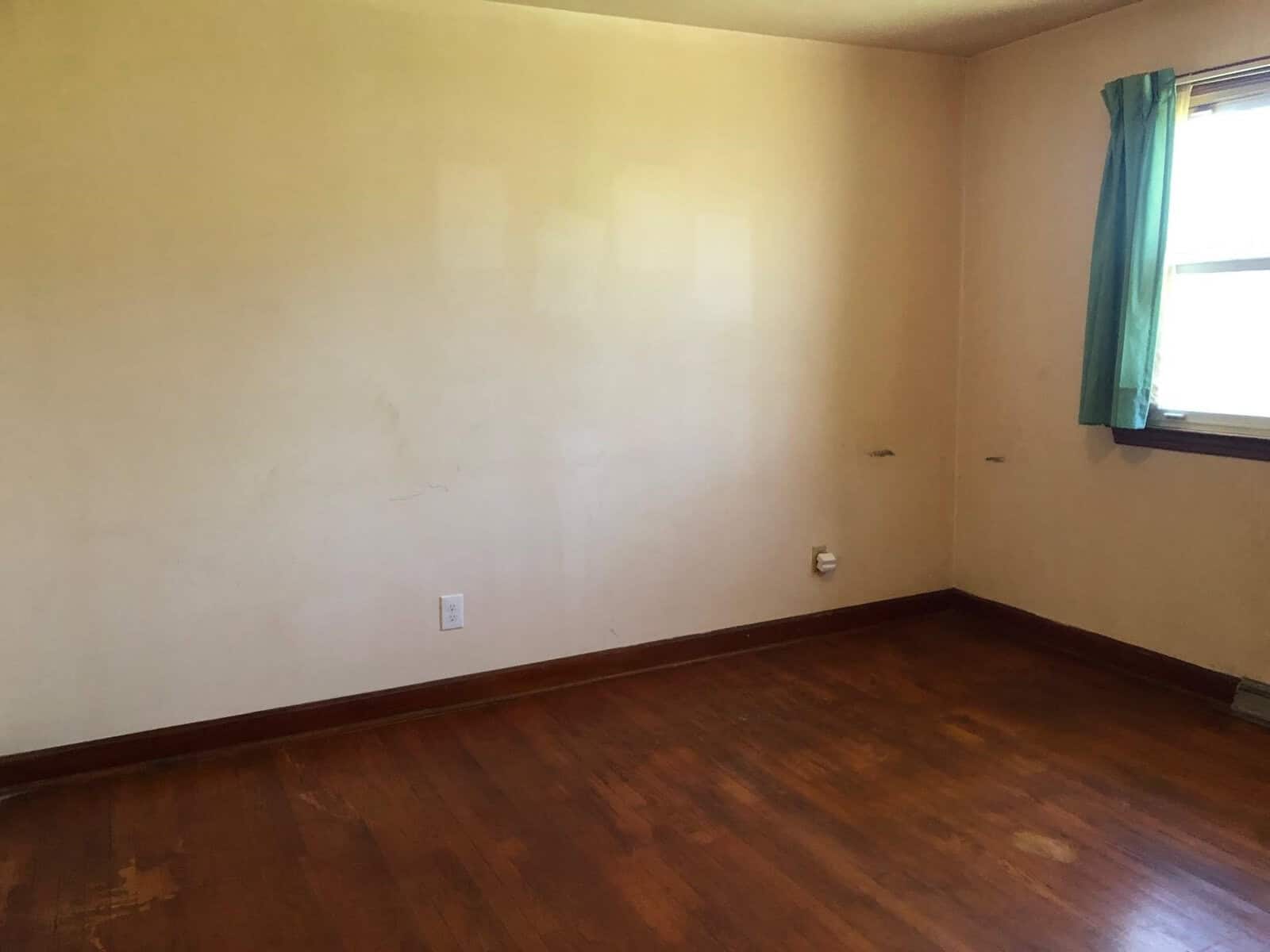 An empty room with hardwood flooring, off-white walls that show some signs of wear, and a single window with a green curtain on the right side.