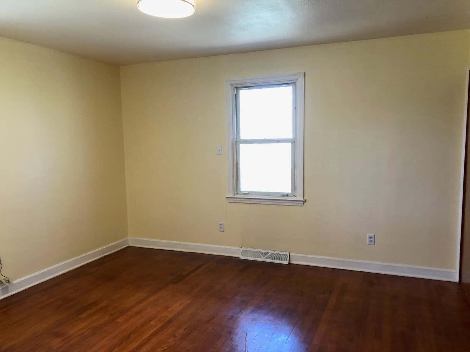 An empty room with beige walls, a single window, and hardwood flooring.