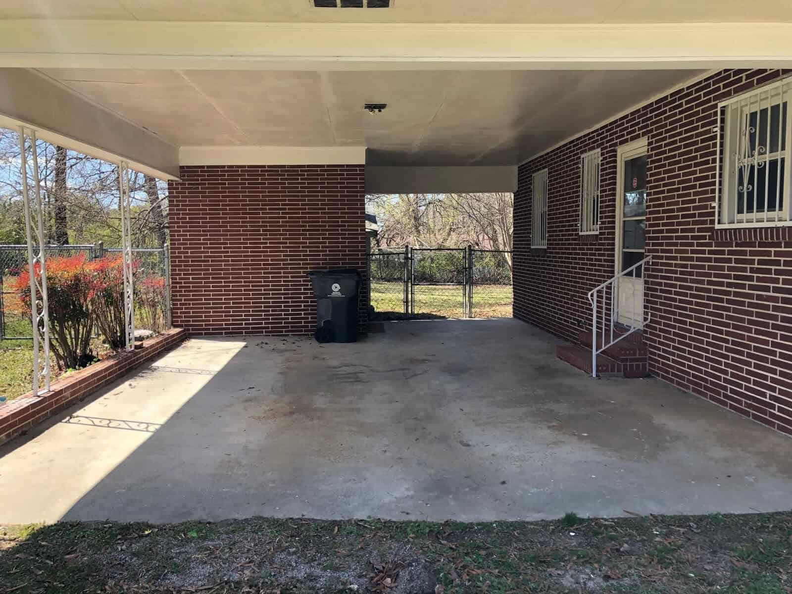 A carport attached to a red brick house with a concrete floor, featuring a black trash bin on the left and a set of stairs leading to a side door.