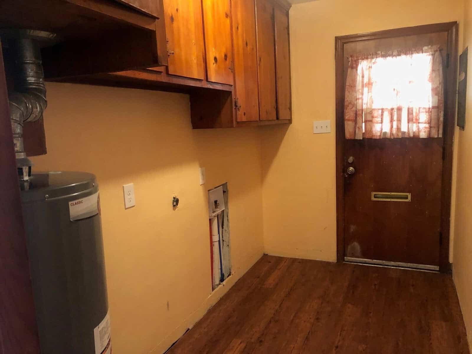 A utilitarian room with wooden flooring features a water heater to the left, unfinished walls with visible plumbing, a wooden cabinet above, an electrical outlet, and a door with glass panes allowing light to enter.