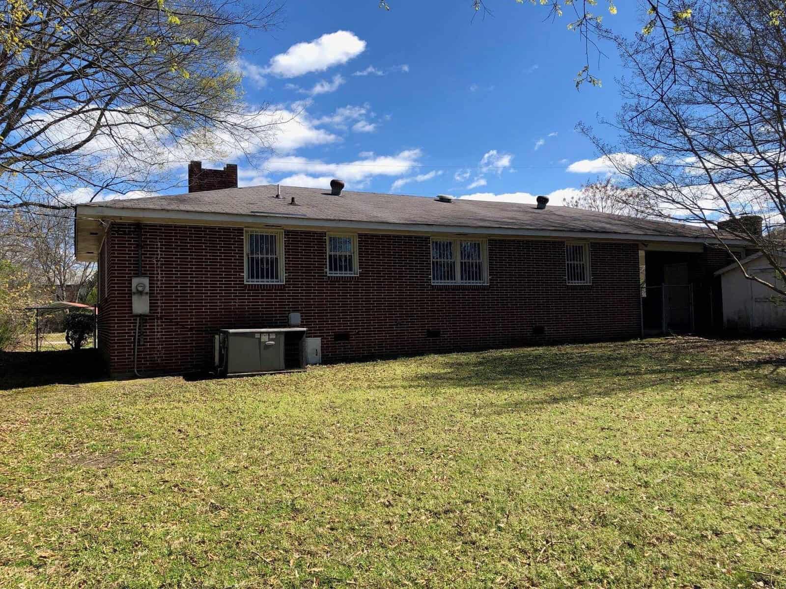 A single-story red brick house with white-trimmed windows, a small chimney, and an attached carport sits under a clear sky with a sparsely treed lawn in the foreground.
