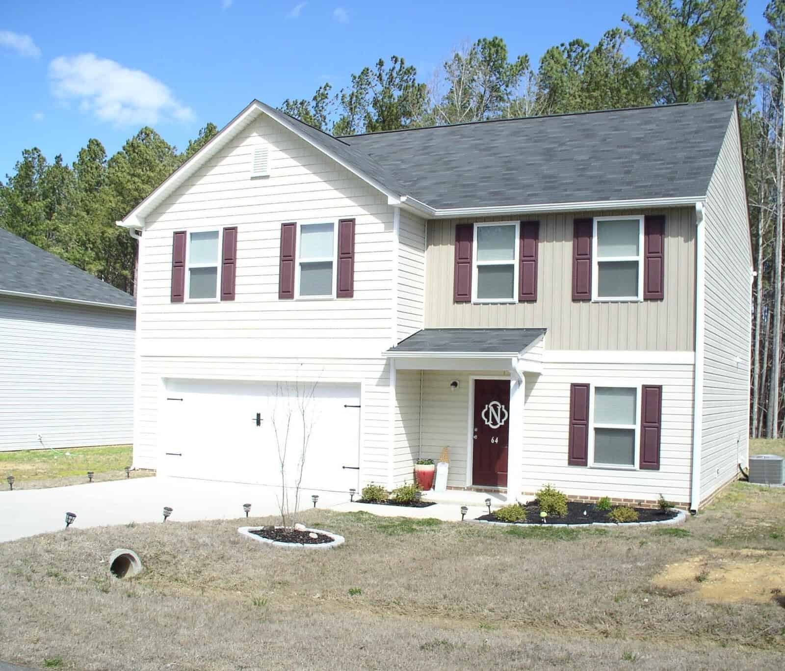 A two-story residential house with white siding and maroon shutters, featuring a garage, a front door with a decorative glass panel, and a small landscaped area by the entrance.