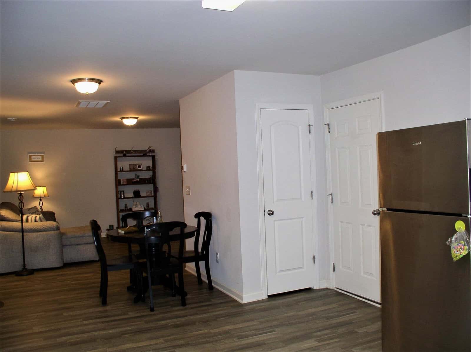 A modern living space featuring a dining area with a black table and chairs, a comfortable seating area with a couch and a side table with a lamp, and a kitchen area with a stainless steel refrigerator. the room has recessed lighting and neutral wall colors.