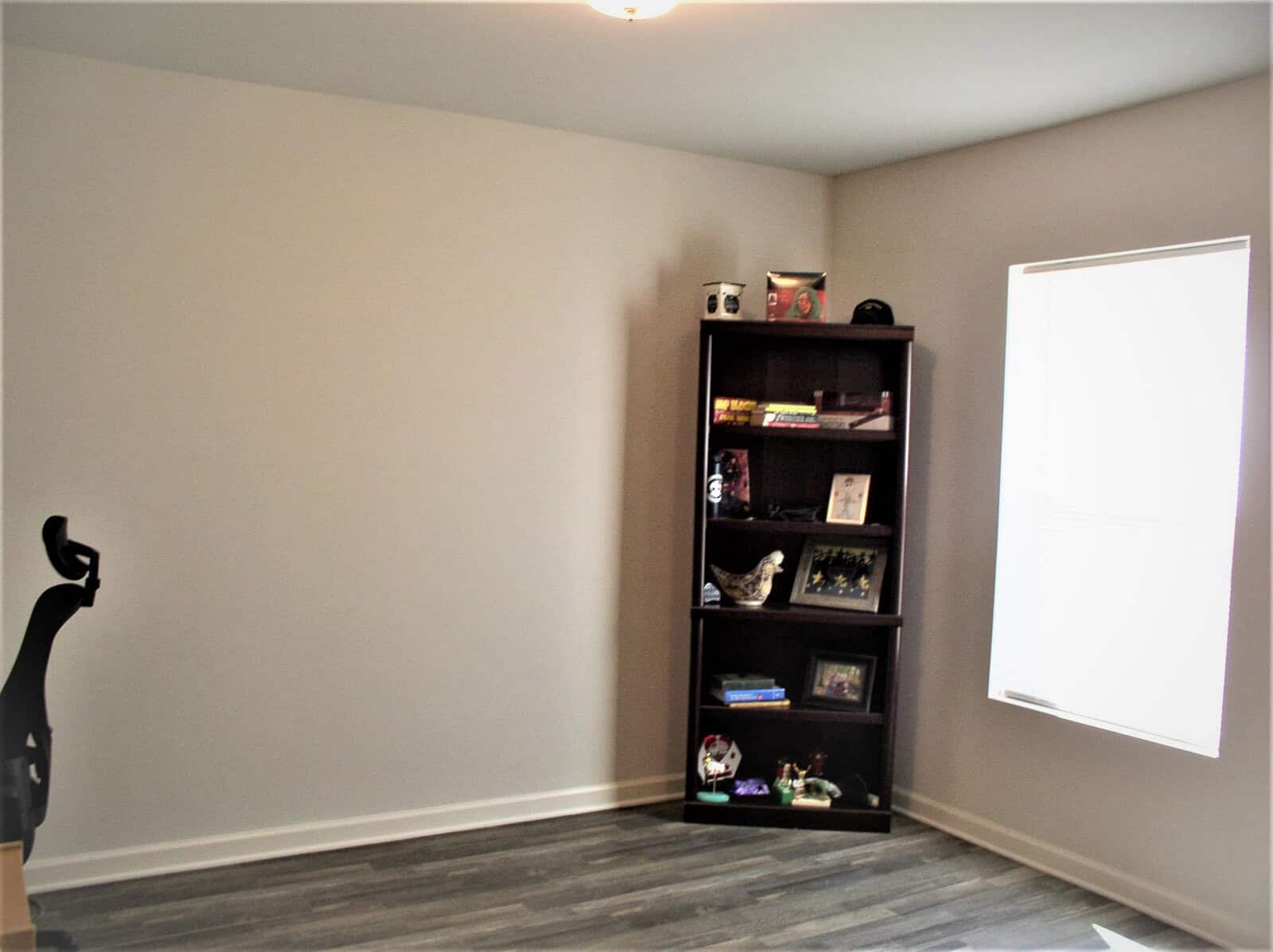 A sparsely furnished room with grey walls and flooring, featuring a tall dark bookshelf filled with a few decorative items and books, next to a large, closed window with a drawn white blind.
