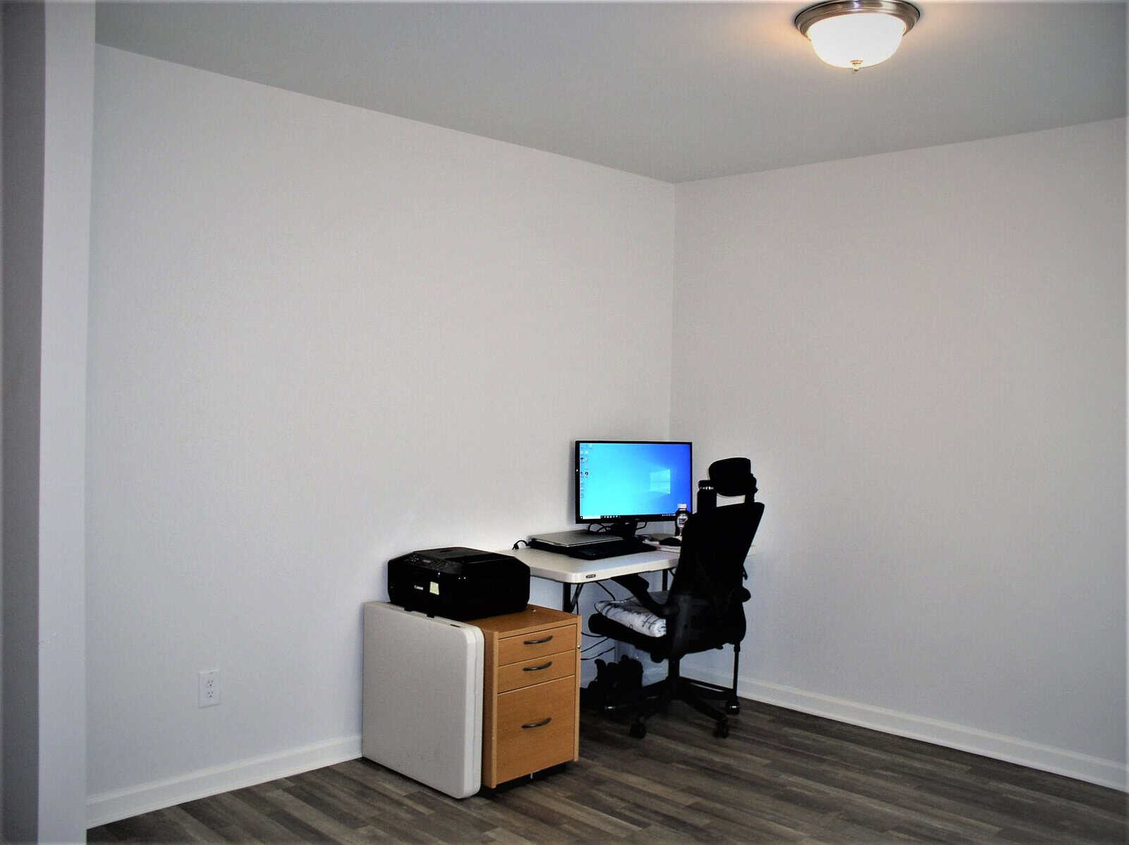 A small, minimally furnished home office with a single chair, desk, computer setup, and printer in a room with white walls and laminate flooring.
