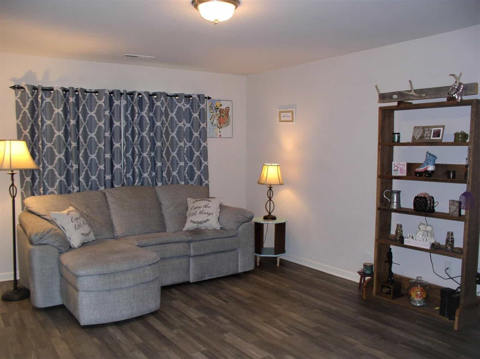 A cozy living room featuring a sectional sofa with decorative pillows, patterned curtains, two standing lamps, and a wooden shelf with various ornaments and framed pictures.