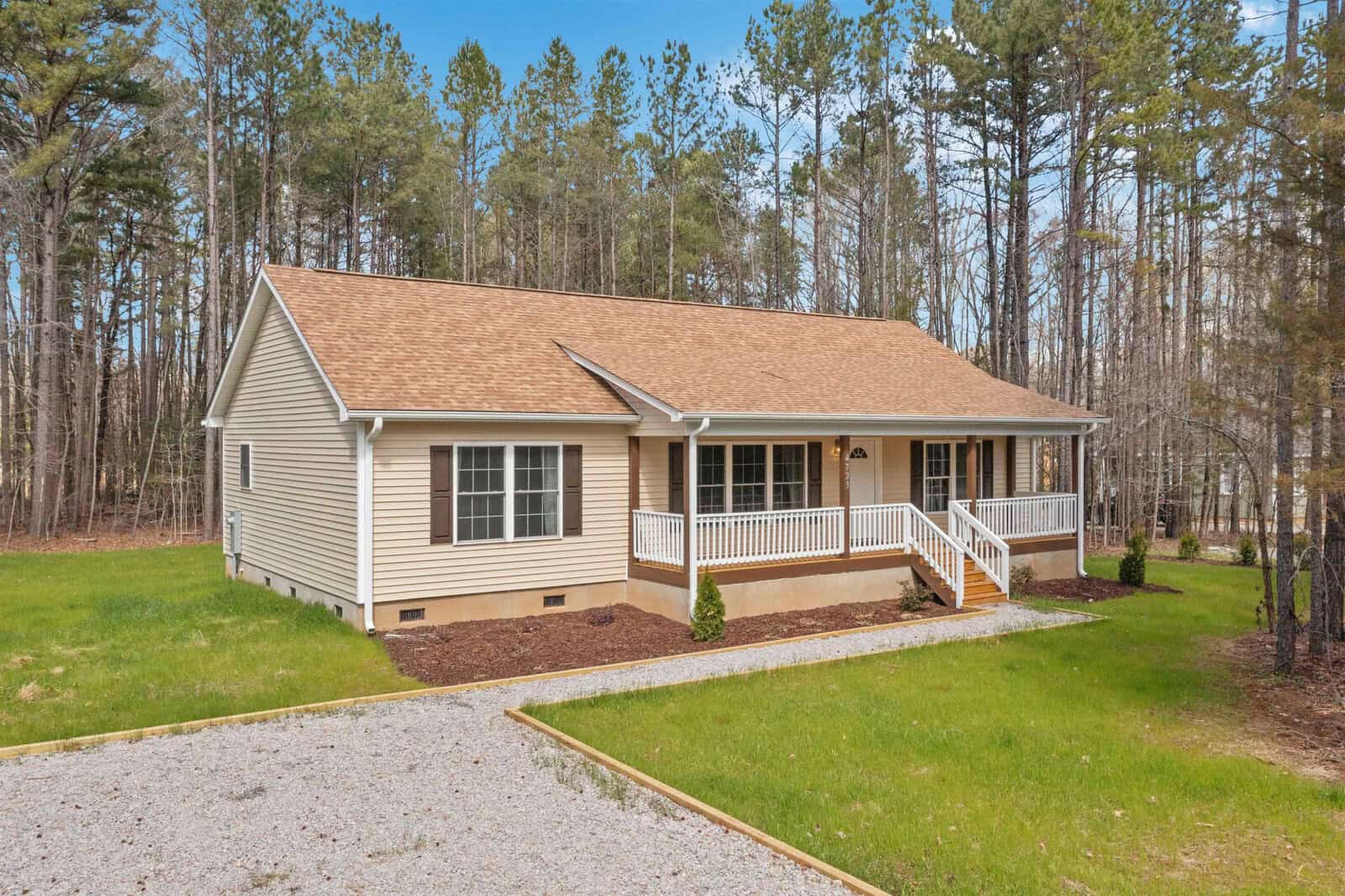 A single-story house with beige siding and a white front porch, set against a backdrop of tall pine trees with a gravel driveway in the foreground.