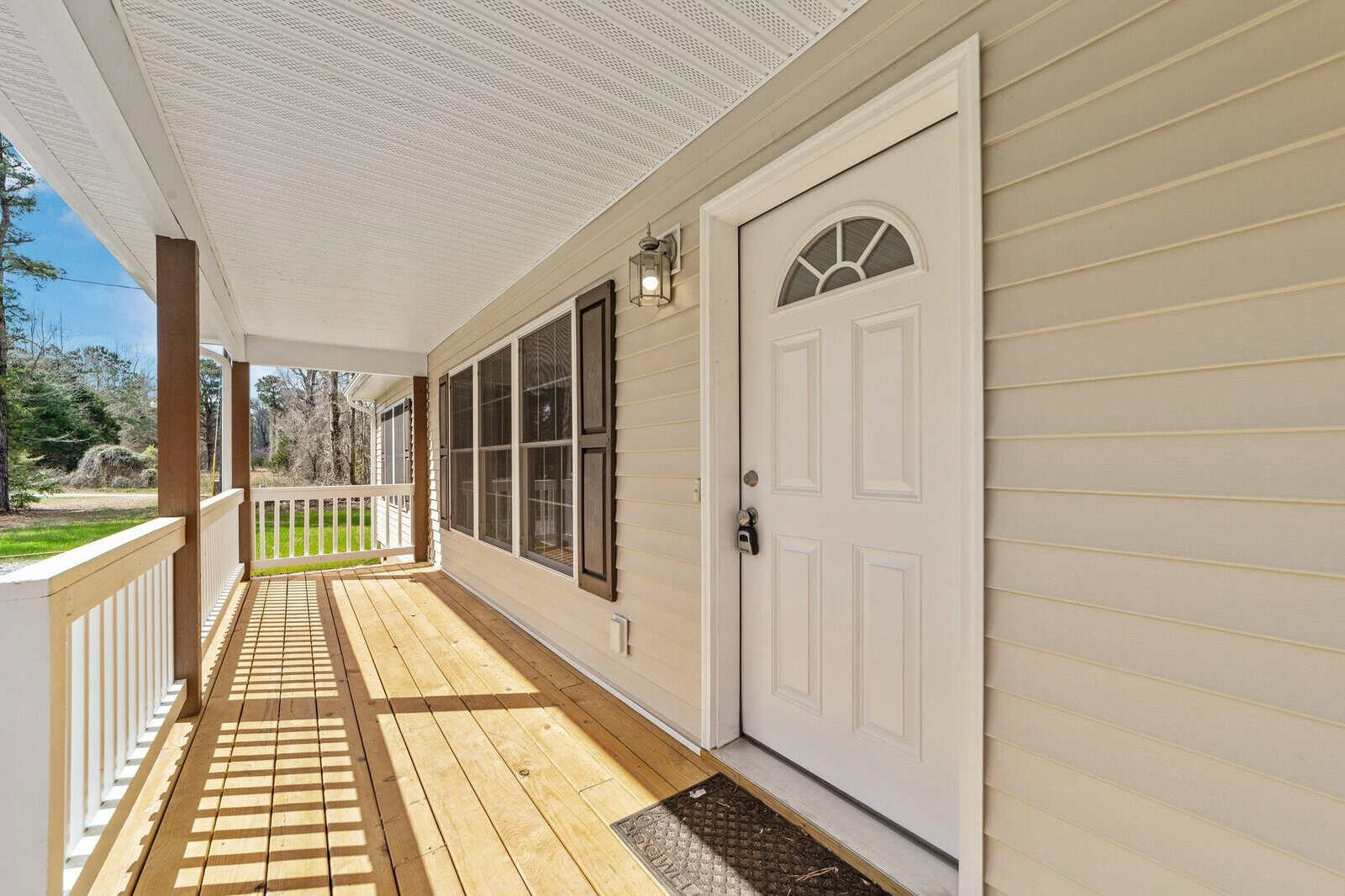 A sunny view of a home's front porch with a white front door and sidelights, featuring wooden flooring, white siding, and a porch ceiling, including an outdoor lantern-style light fixture.