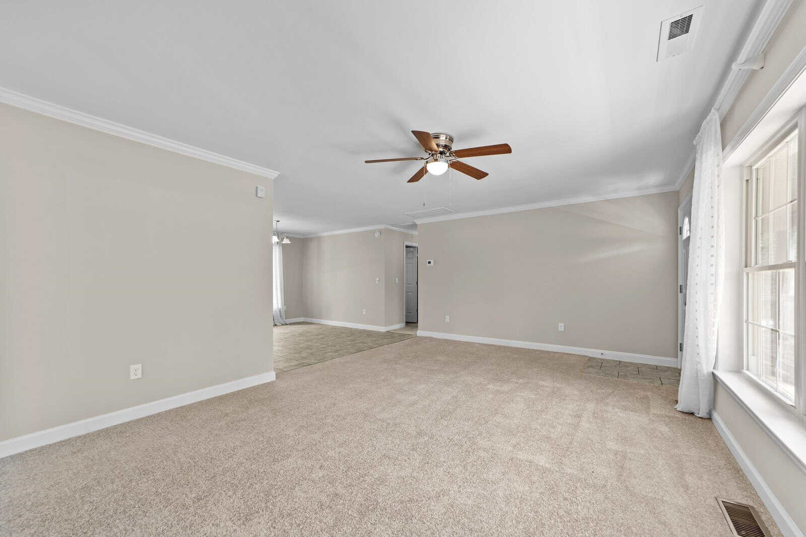 A spacious, empty room with beige carpeting, white walls, a ceiling fan, and multiple windows, suggesting a well-lit area.