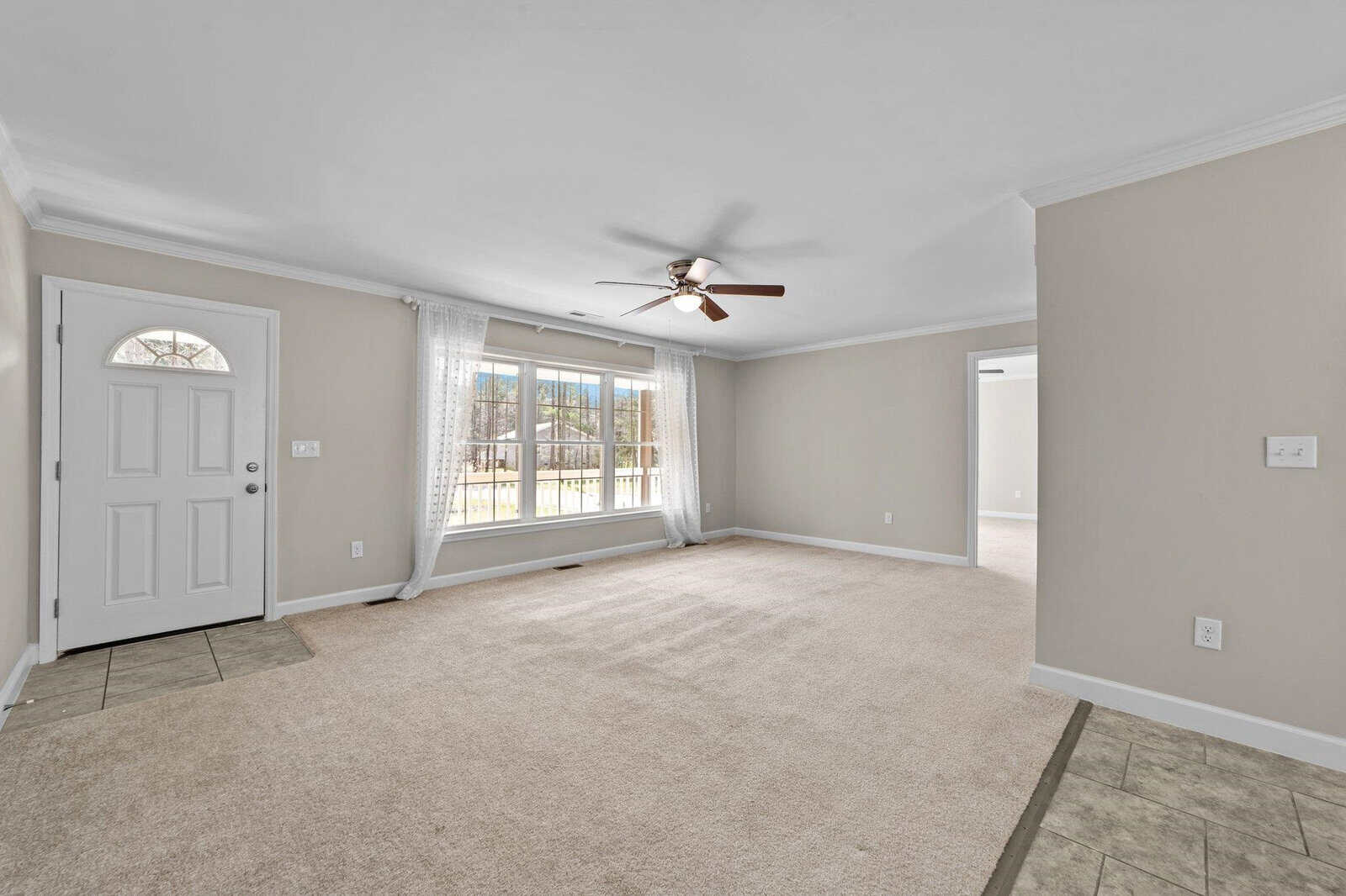 An empty room with beige carpeting, a ceiling fan, white walls with one accent wall, and large windows with curtains next to a white front door.