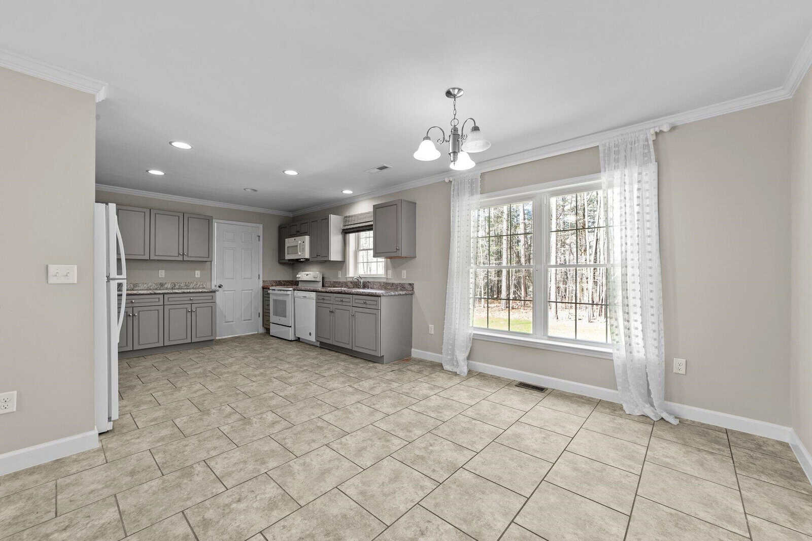 A spacious, empty kitchen and dining area with tile flooring, grey cabinets, white walls, and ample natural light from large windows draped with sheer curtains.
