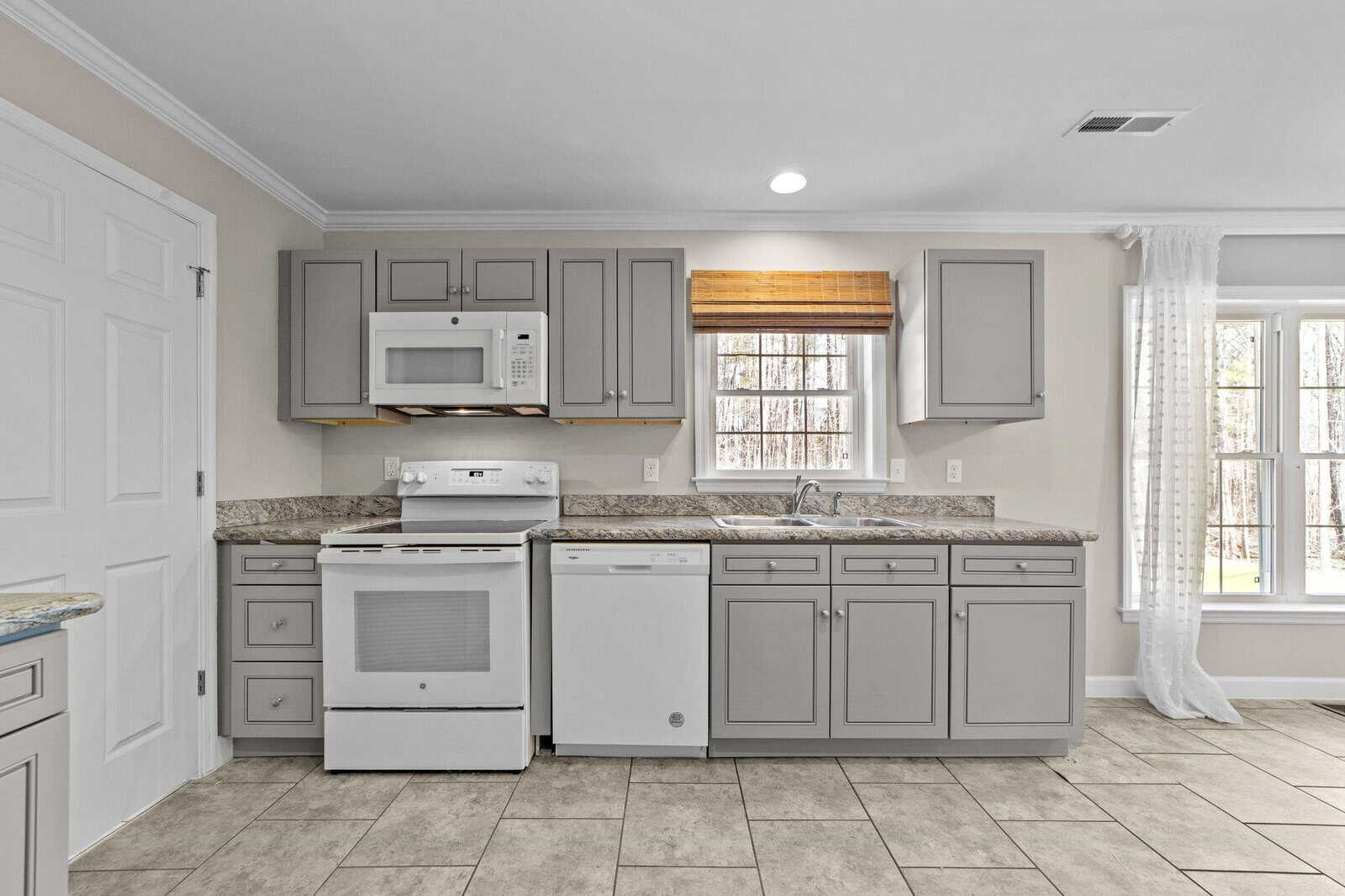 A clean and empty kitchen interior featuring gray cabinets, white appliances, granite countertops, tiled flooring, and a window with a brown blind.