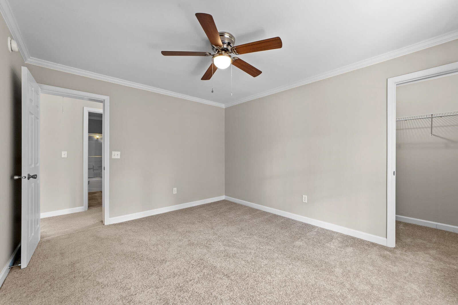 A vacant room with beige walls and carpet flooring featuring an illuminated ceiling fan and open doors leading to other rooms.