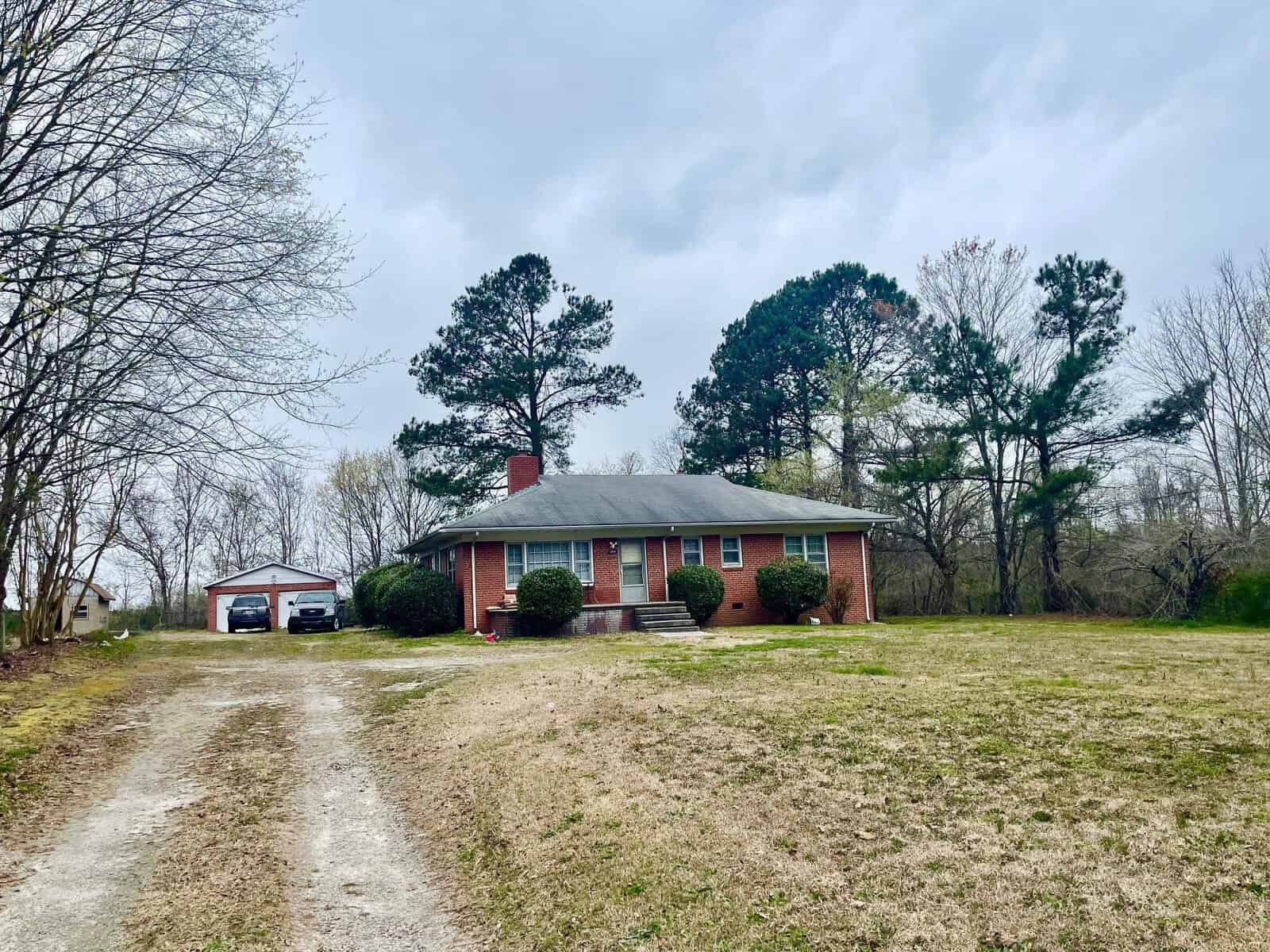 A single-story red brick house with a gable roof and two front steps, flanked by leafless trees and a green lawn, with an overcast sky overhead and a separate garage structure in the background on the right.
