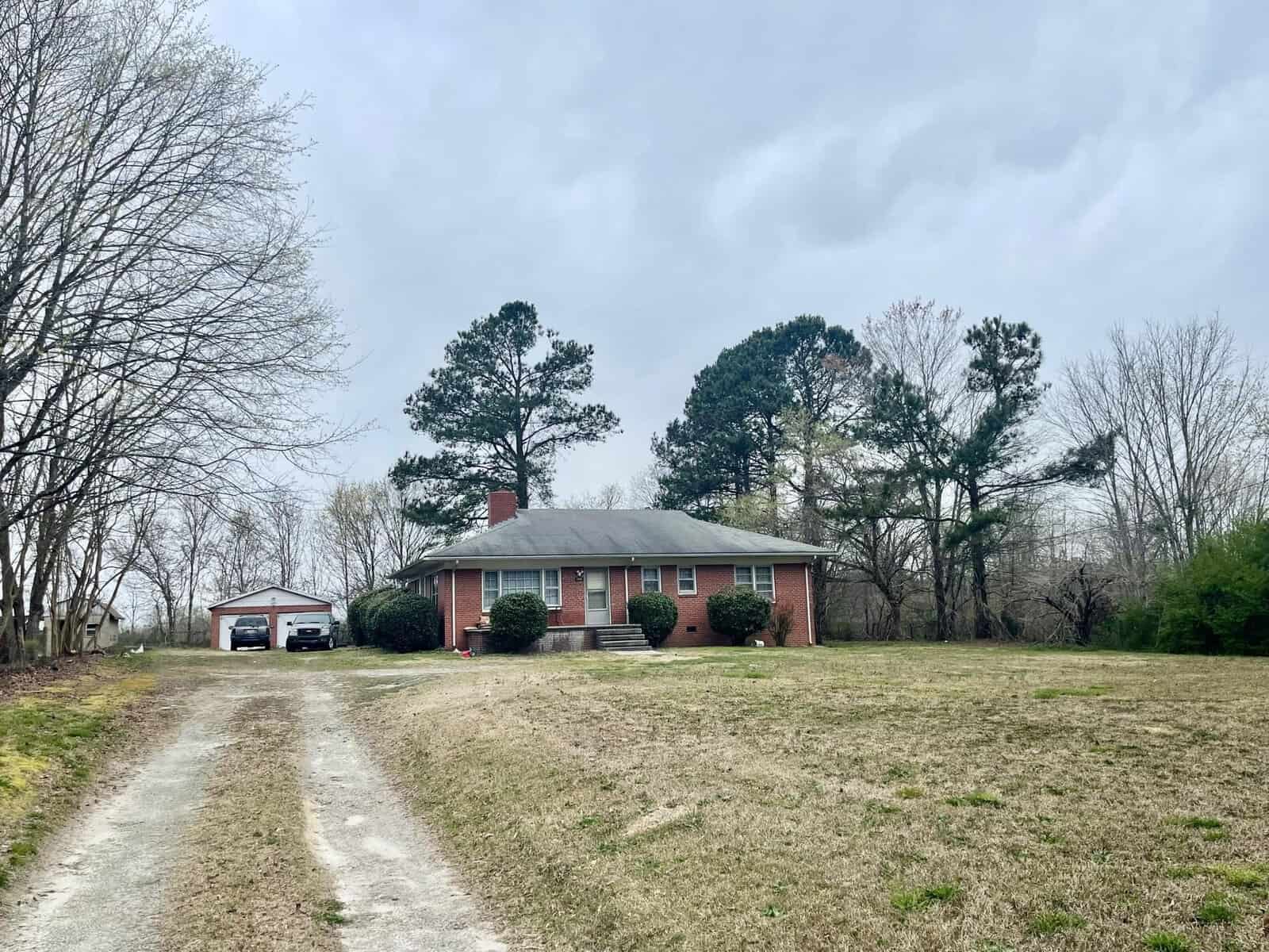 A single-story red brick house with a white door and two white-framed windows, a detached garage to the left, and a bare tree-lined driveway leading up to the house, neighbored by a grassy front yard, under an overcast sky.