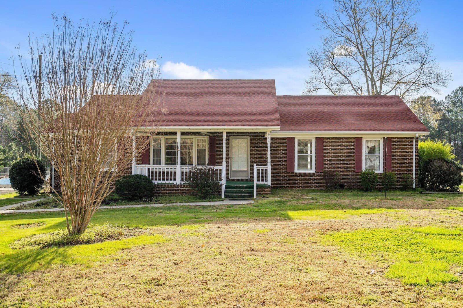 A single-story brick house with a red roof and a small front porch, situated on a lawn with scattered trees and shrubs under a clear sky.