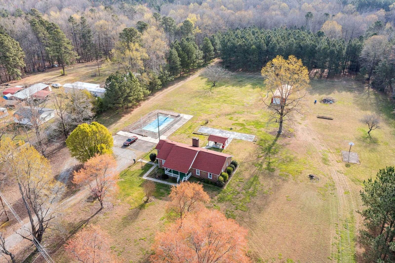 Aerial view of a red brick house with a backyard swimming pool surrounded by trees and adjacent open land.