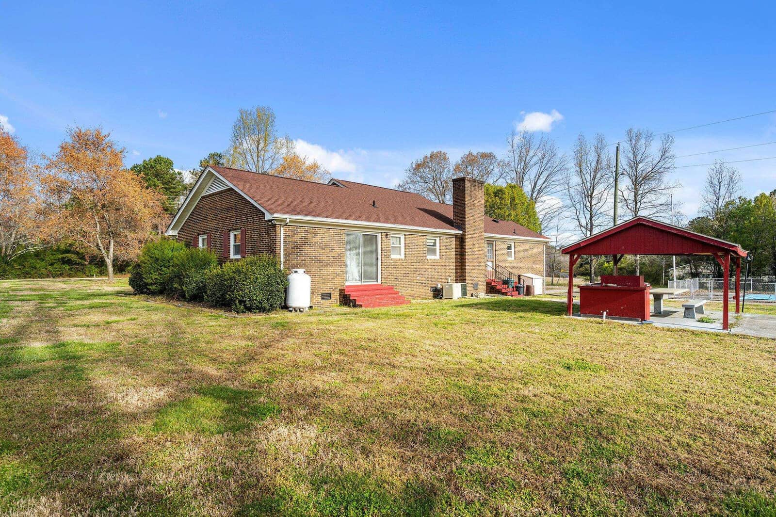 A single-story brick house with a chimney, featuring a large backyard with a swimming pool under a red canopy and surrounded by trees, under a clear blue sky.