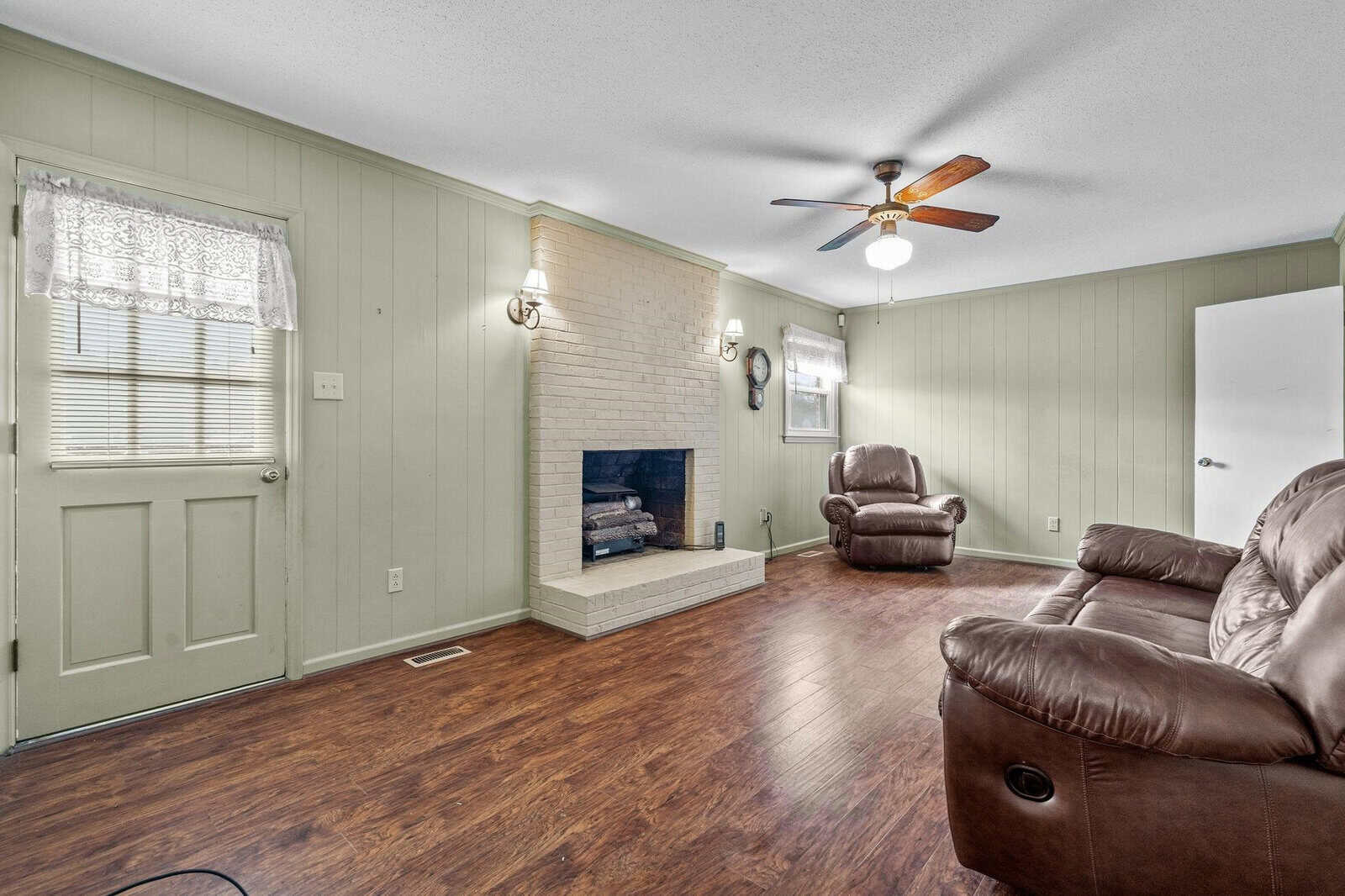 Spacious living room featuring a hardwood floor, a white brick fireplace, paneled walls painted in a light green, two dark leather armchairs, and a ceiling fan.