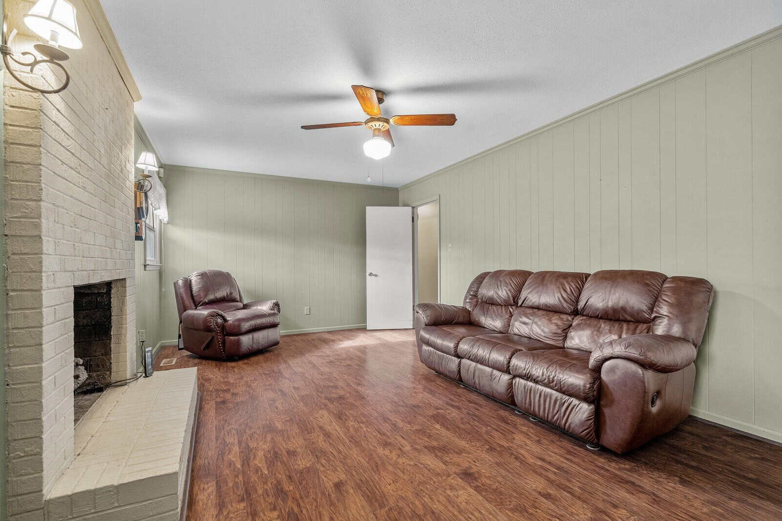 A sparsely furnished living room with a brown leather sofa and recliner, featuring a brick fireplace, hardwood floors, and a ceiling fan.
