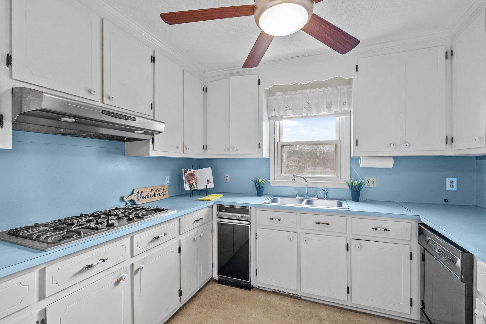 A bright kitchen featuring white cabinetry, blue countertops, and walls partially painted in blue, with a window above the sink allowing natural light, and a ceiling fan above.