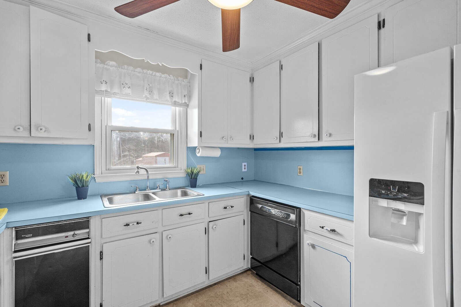 A bright kitchen interior with white cabinetry, light blue walls, a double sink, dishwasher, and a fridge, highlighted by natural light from two windows.